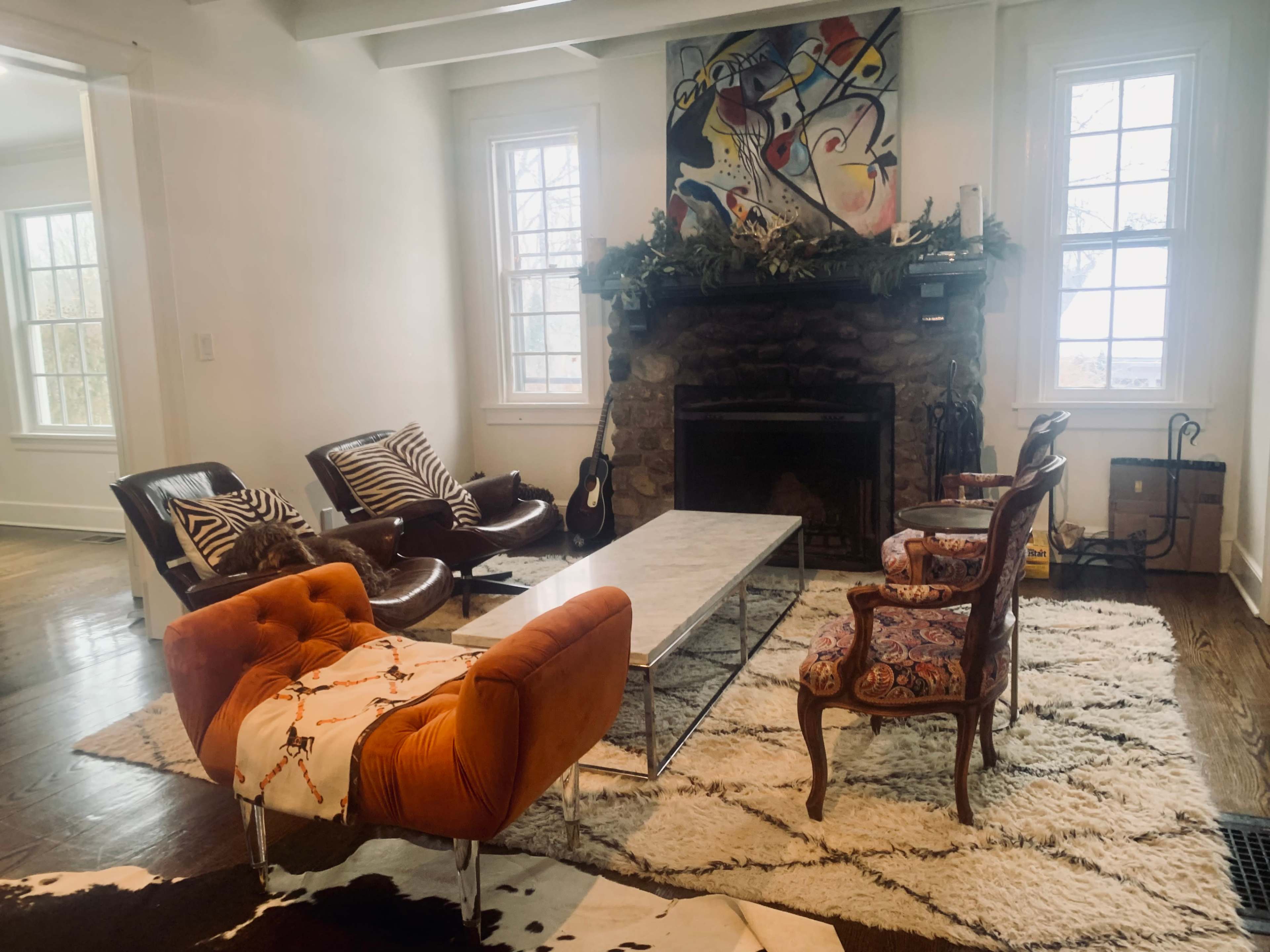 The room features a stone fireplace with a mantel adorned with decorations, surrounded by various seating options, including two armchairs, a modern couch, and a coffee table on a patterned rug.