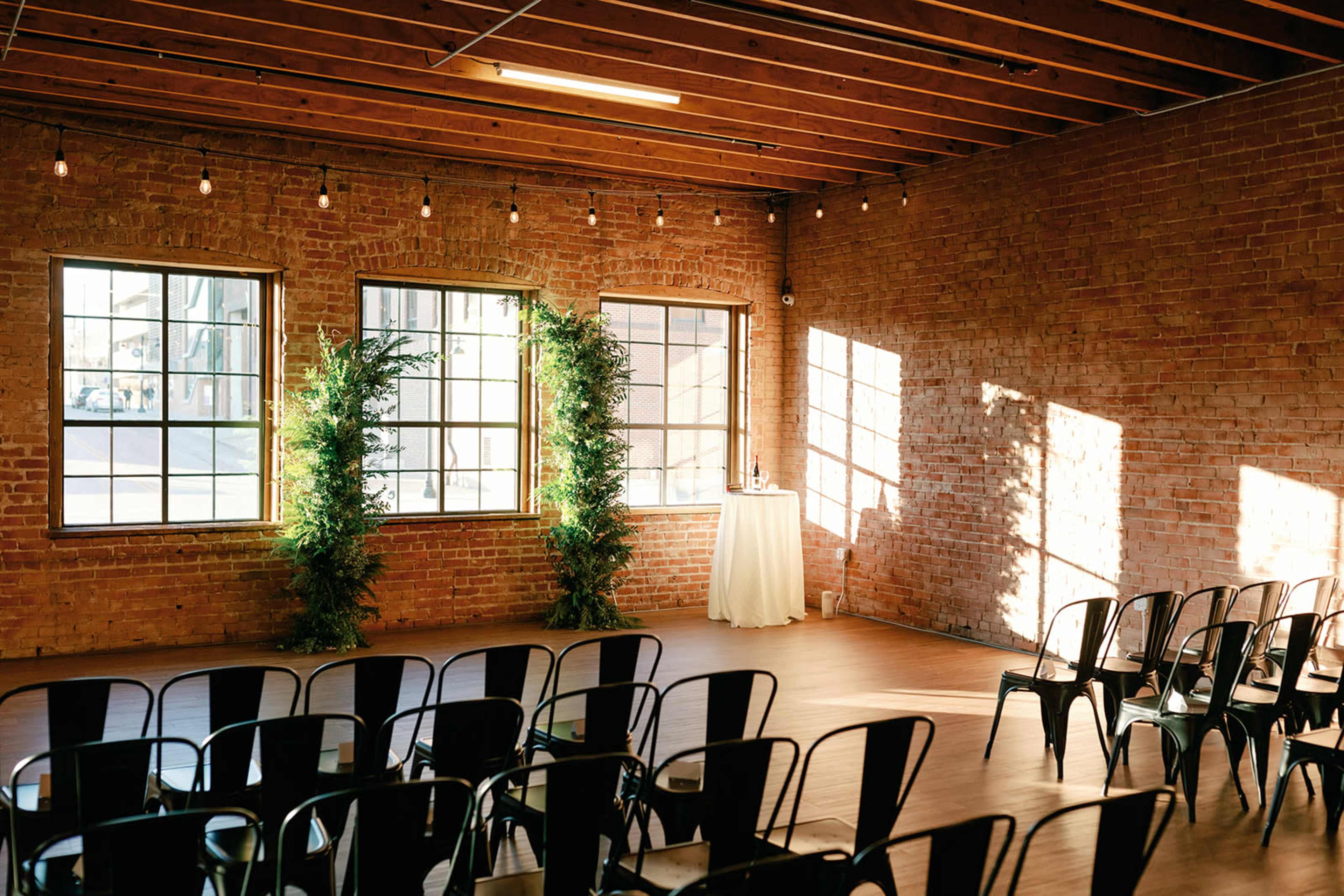 The image shows a brick-walled venue with rows of black chairs facing a small table adorned with greenery and set against large windows that let in natural light.