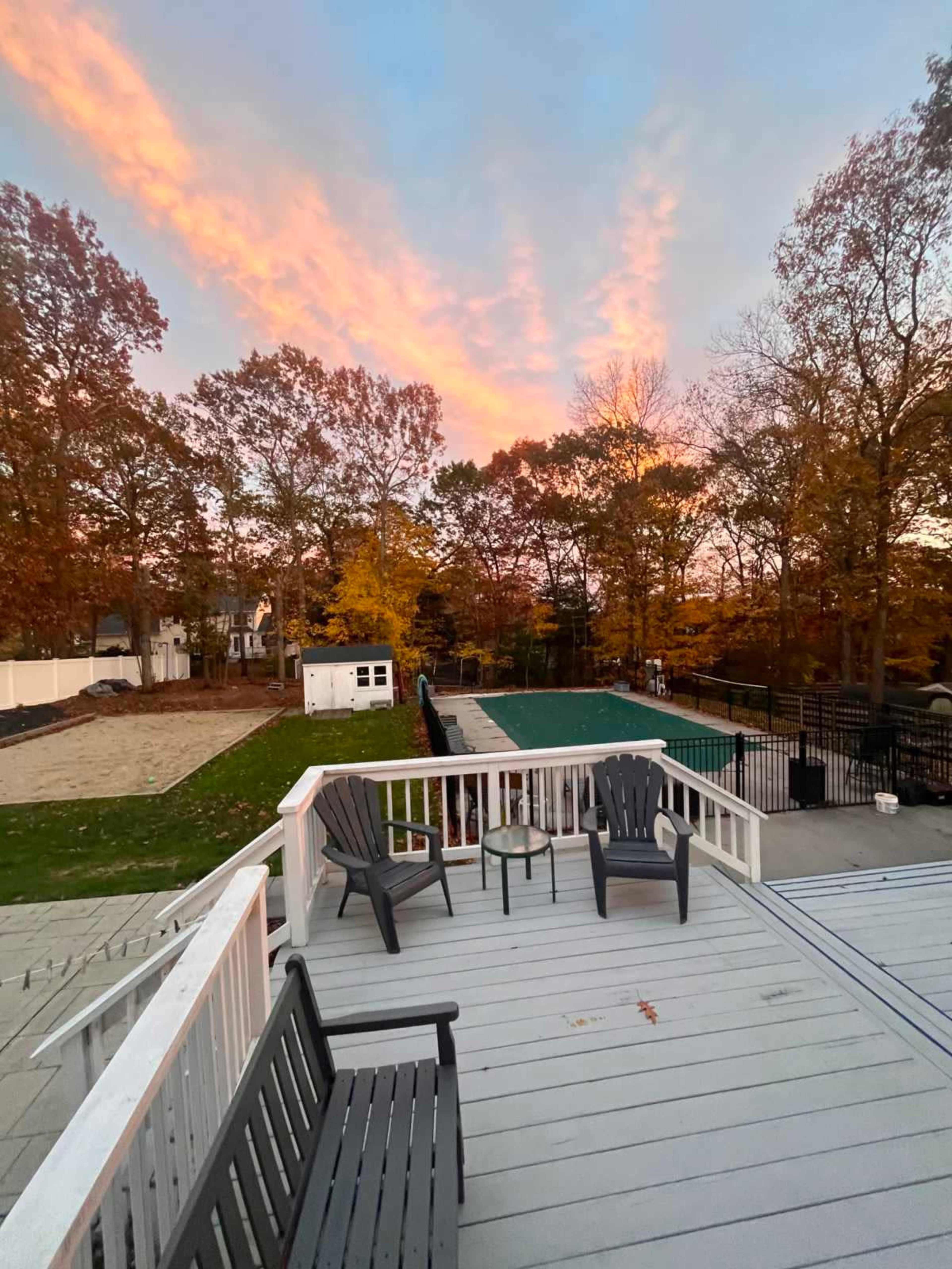 A deck with chairs overlooks a backyard featuring a swimming pool, sand area, and trees under a colorful sky at sunset.