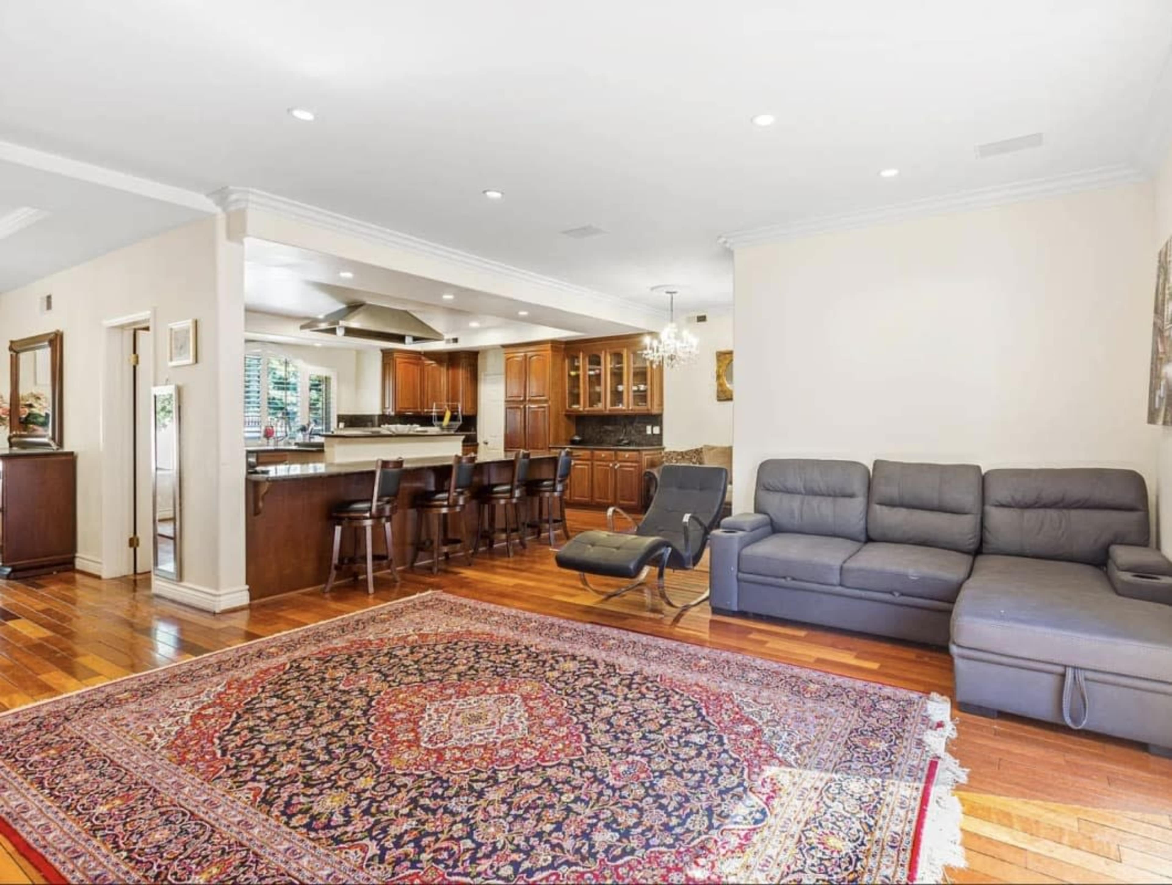 The image shows a spacious living room featuring a large area rug, a sectional couch, and an open kitchen with wooden cabinets in the background.