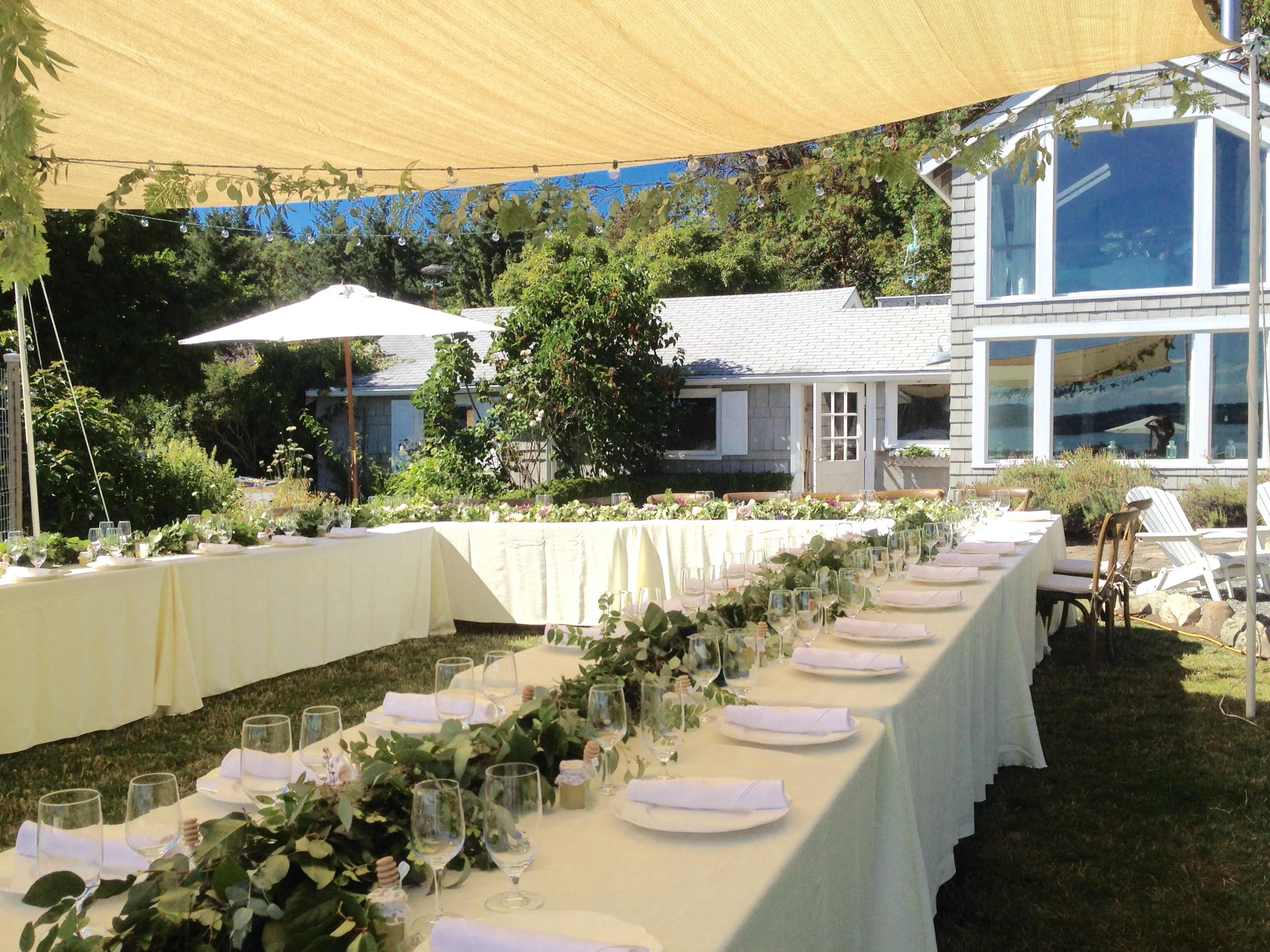 A long banquet table is set outdoors under a yellow canopy, surrounded by greenery and a spacious house in the background.