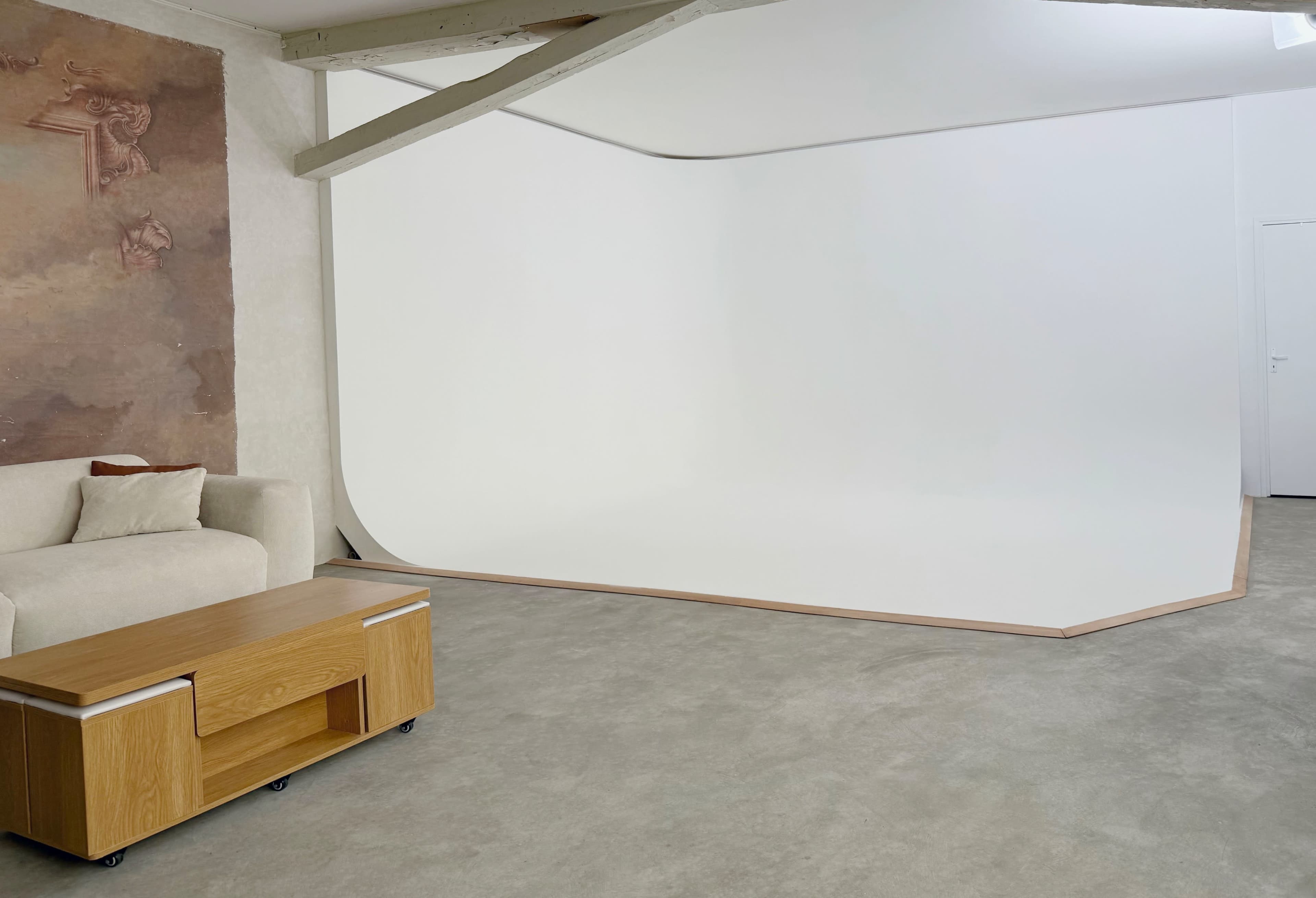 The image shows a spacious, minimalist studio with a white seamless backdrop, a wooden coffee table, and a light-colored couch against a textured wall.