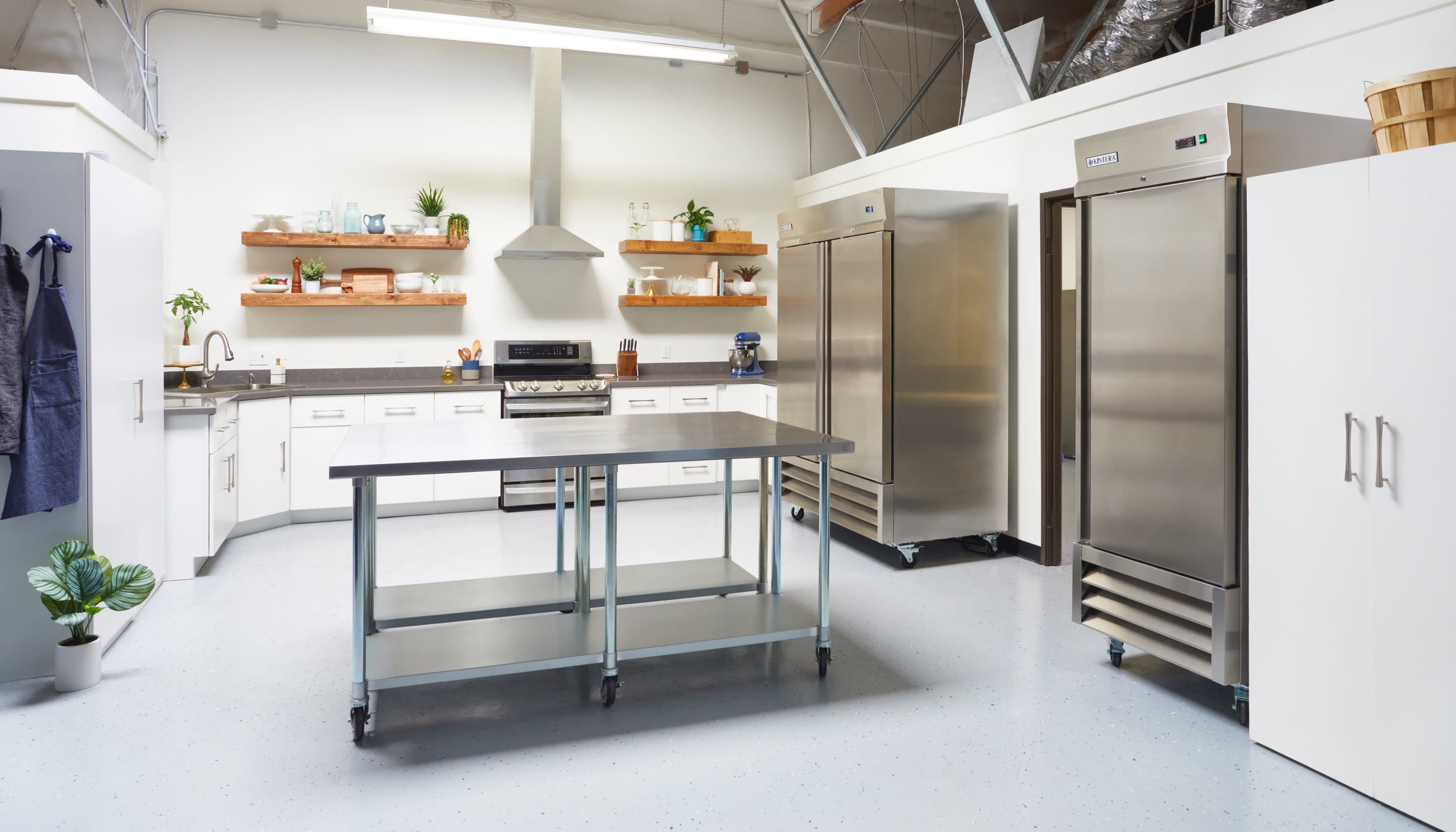 The image shows a spacious kitchen equipped with stainless steel appliances, including two refrigerators, countertops, and shelves with kitchenware.