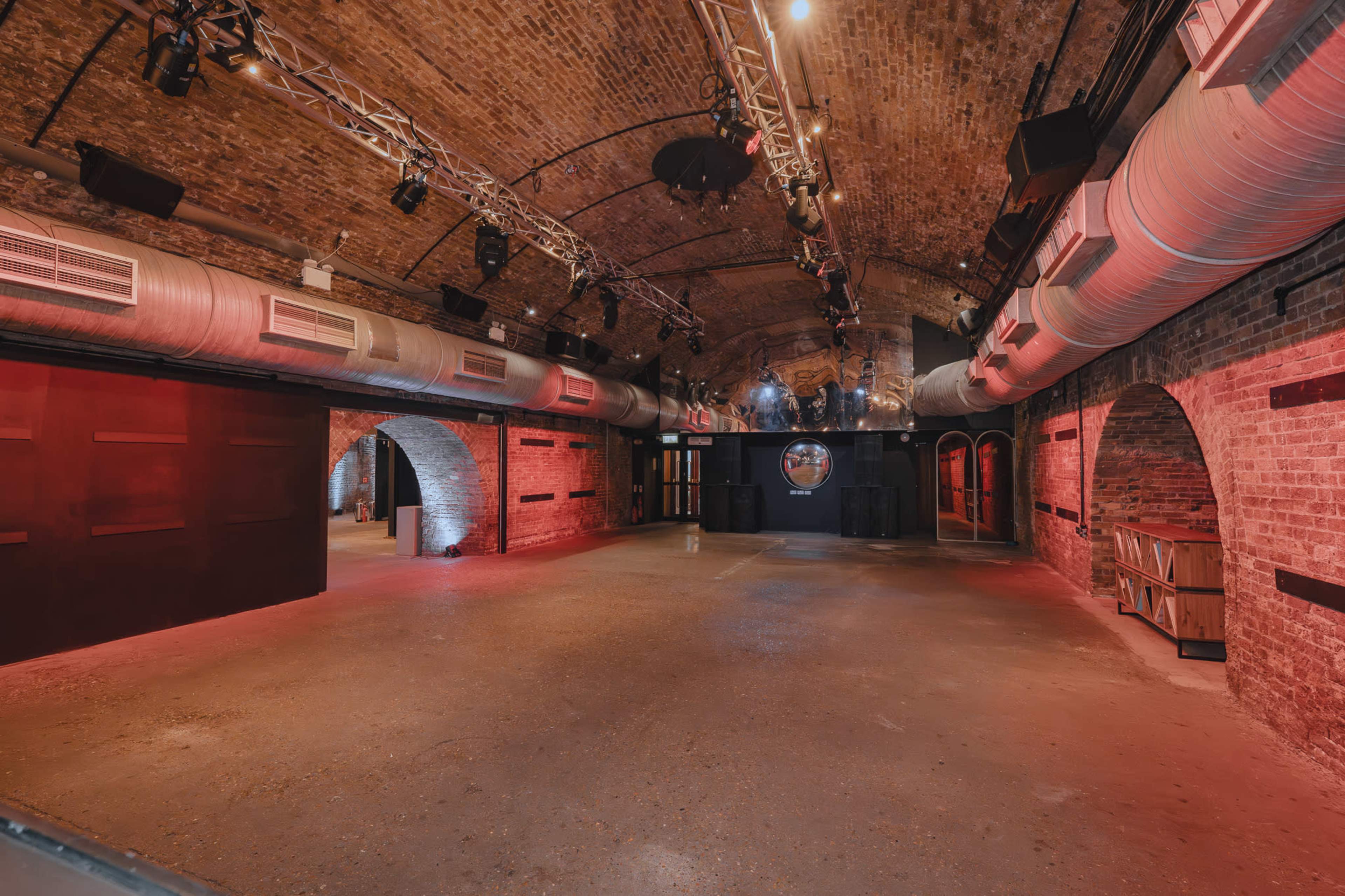 The image shows a spacious, empty event space with exposed brick walls, arched ceilings, and red lighting accents.