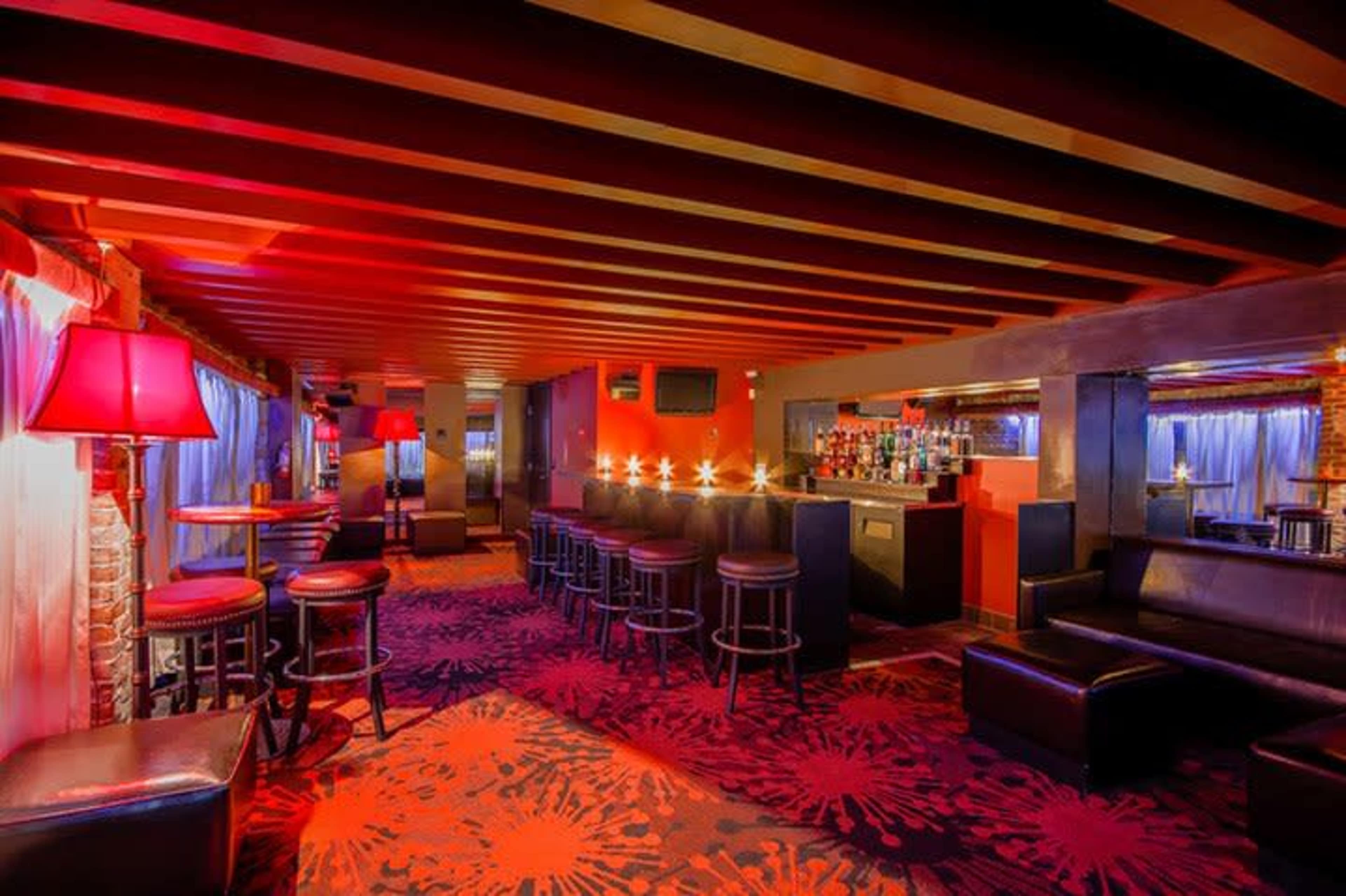 The image shows a dimly lit lounge with red lighting, equipped with bar stools, low tables, and a bar area against one wall.