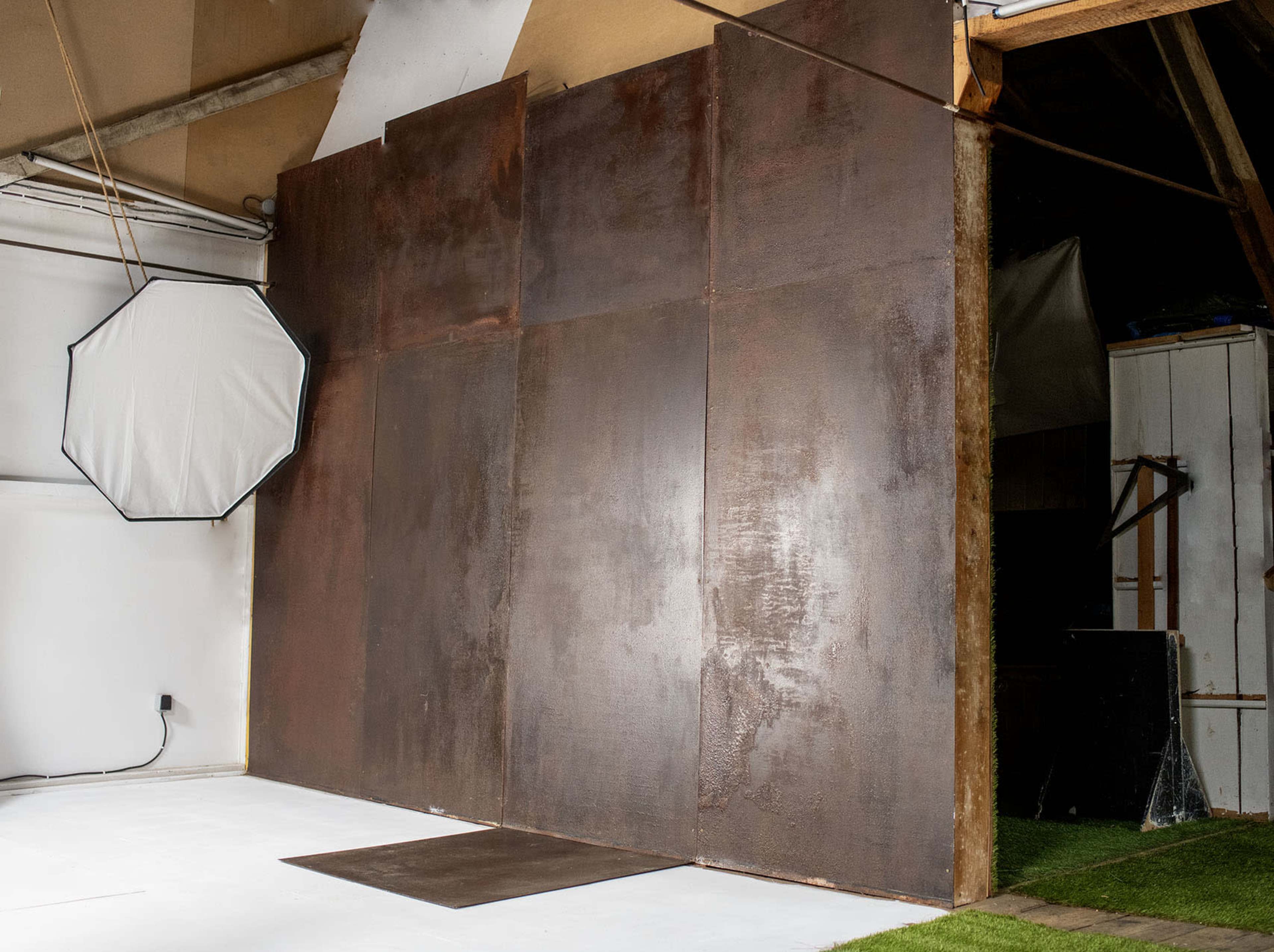 The image shows a studio space featuring a large, rusted metal backdrop with an octagonal softbox light positioned to the left.