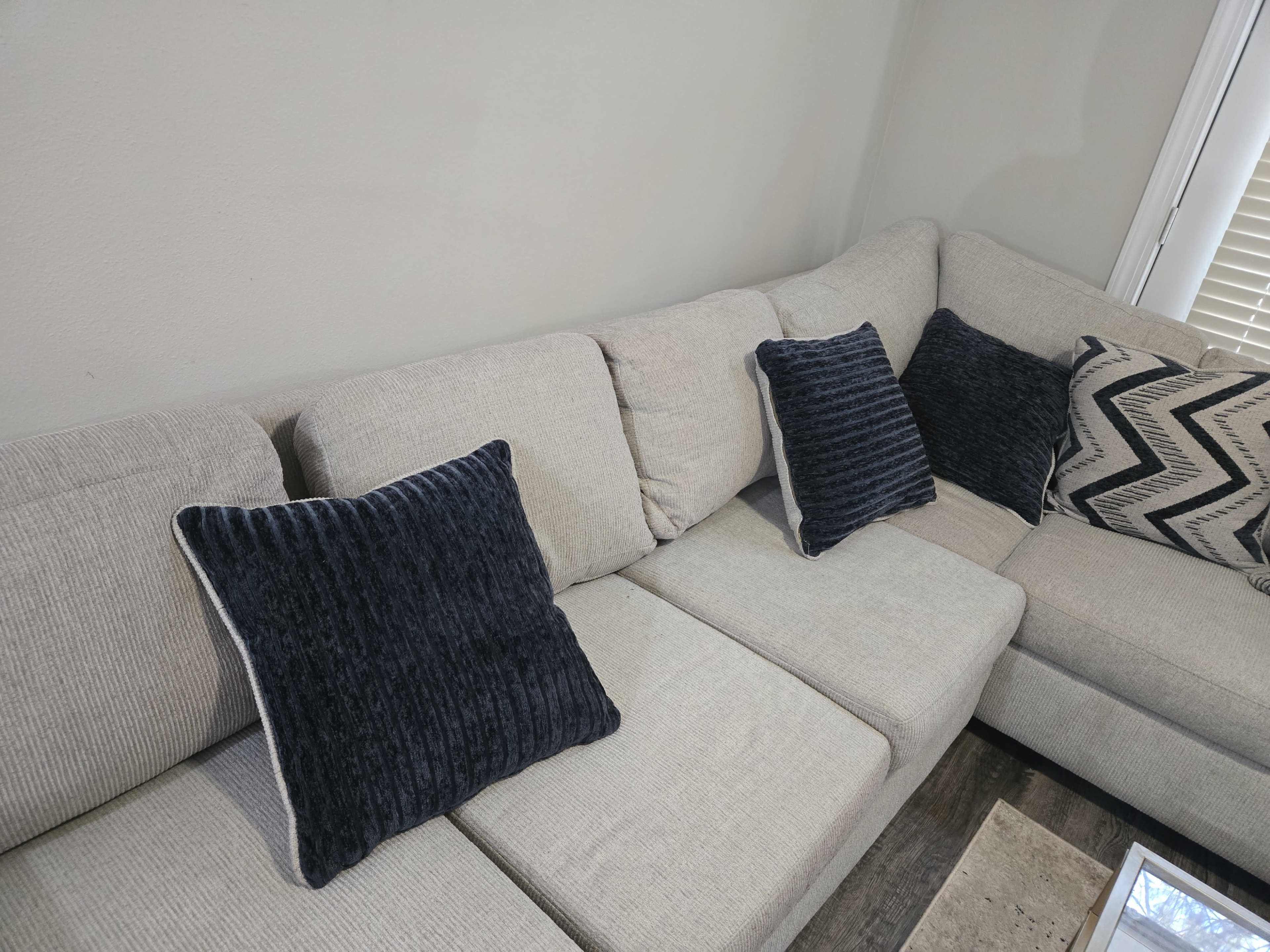 The image shows a light gray sectional sofa with two dark blue textured throw pillows and a zigzag patterned pillow on the corner.