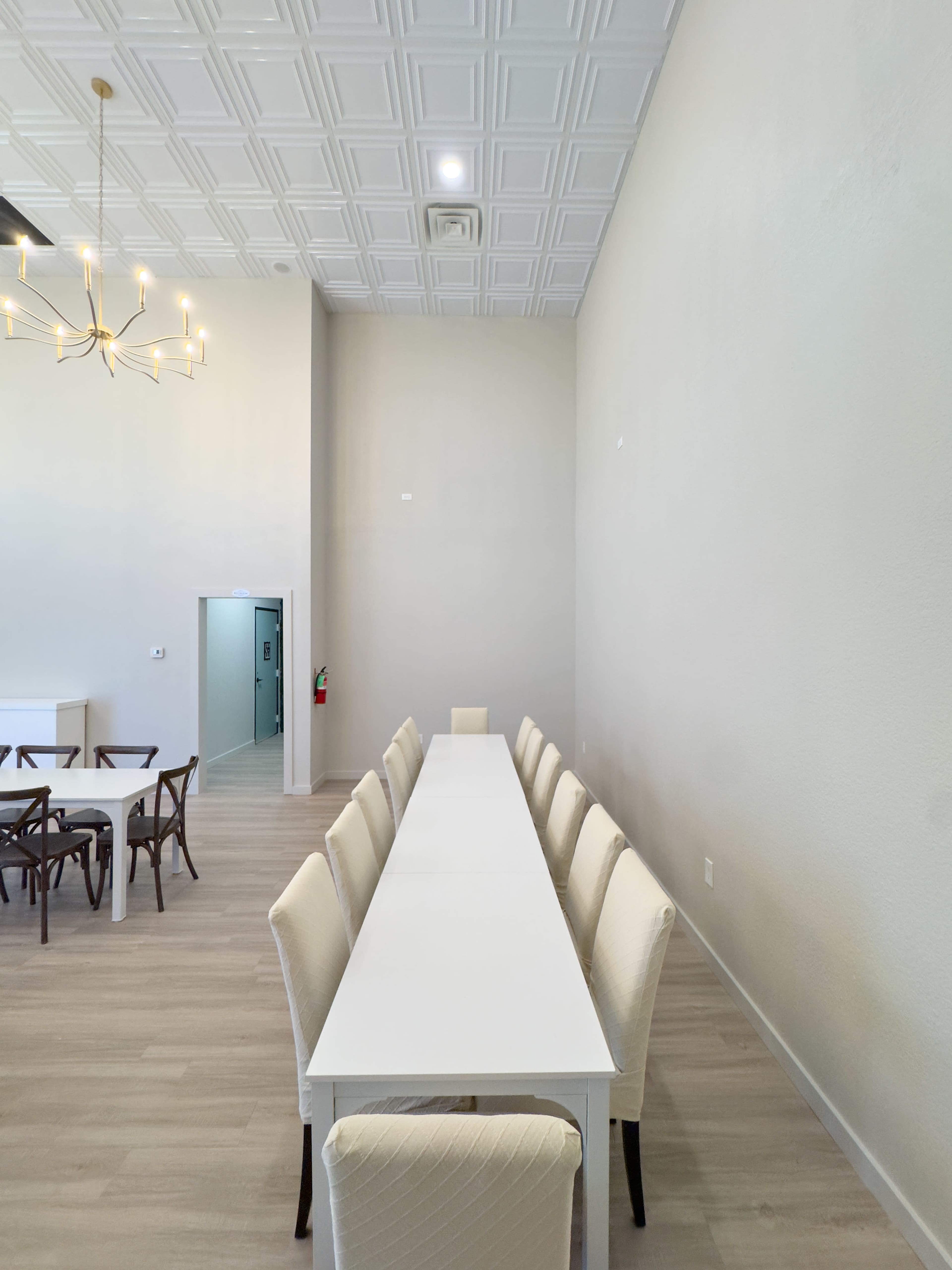 A long white table with beige chairs is positioned against a light-colored wall in a spacious room.