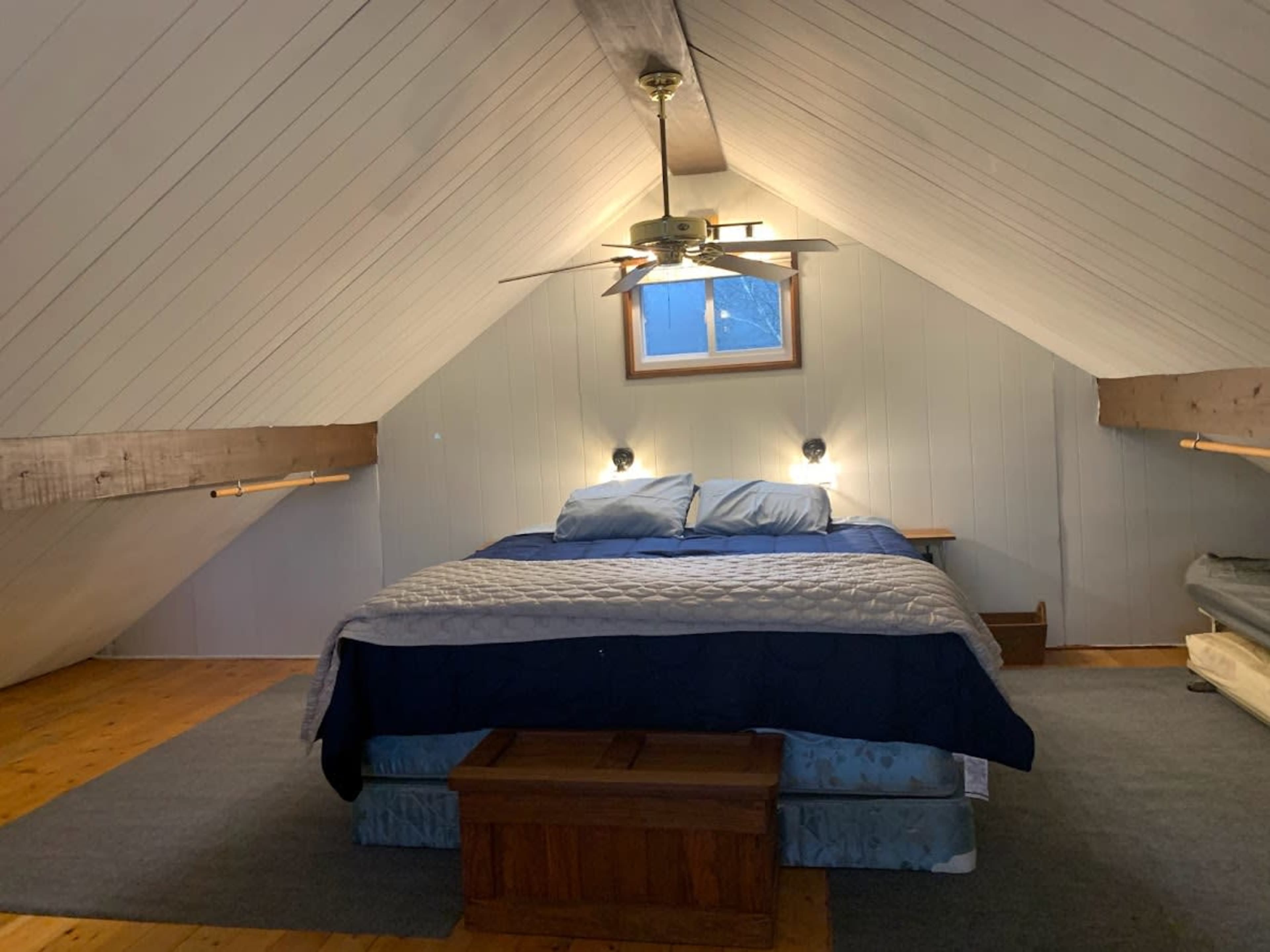 A cozy attic bedroom featuring a large bed with blue bedding, a ceiling fan, and wooden beams along the sloped ceiling.