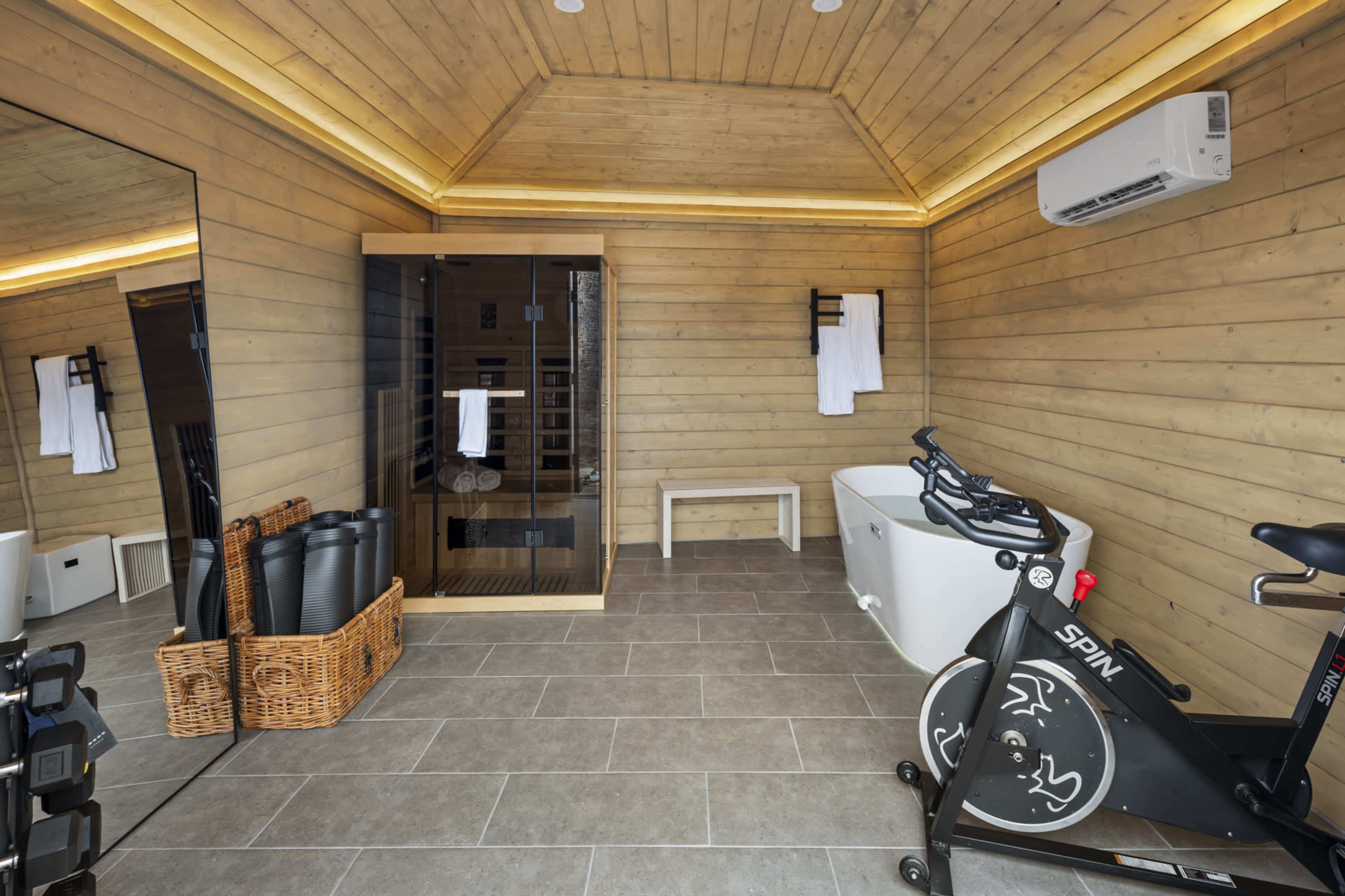 The image shows a modern, wooden-paneled bathroom equipped with a sauna, bathtub, exercise bike, and storage for towels and fitness equipment.