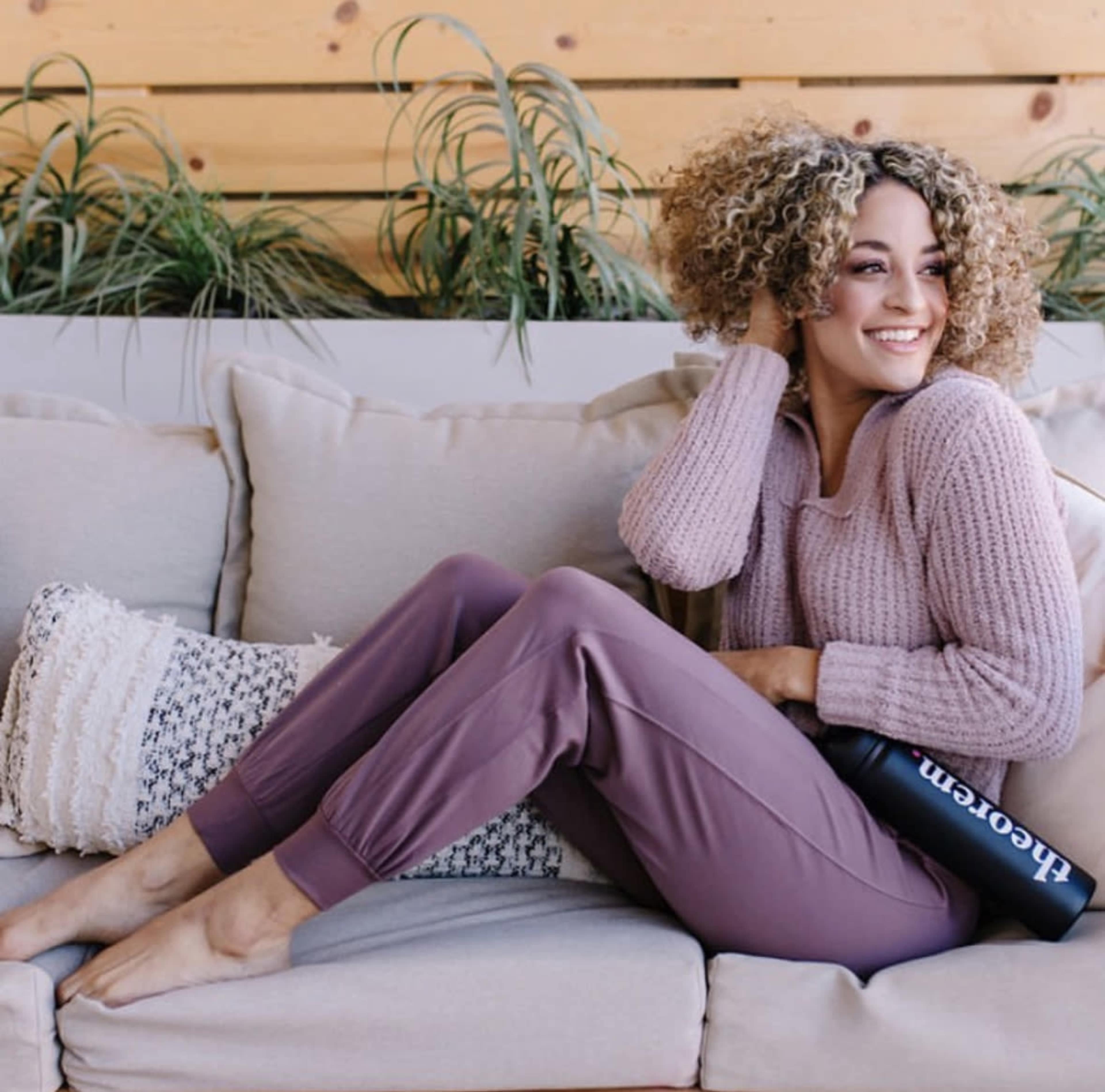 A woman with curly hair sits on a couch wearing a sweater and comfortable pants, holding a black water bottle.