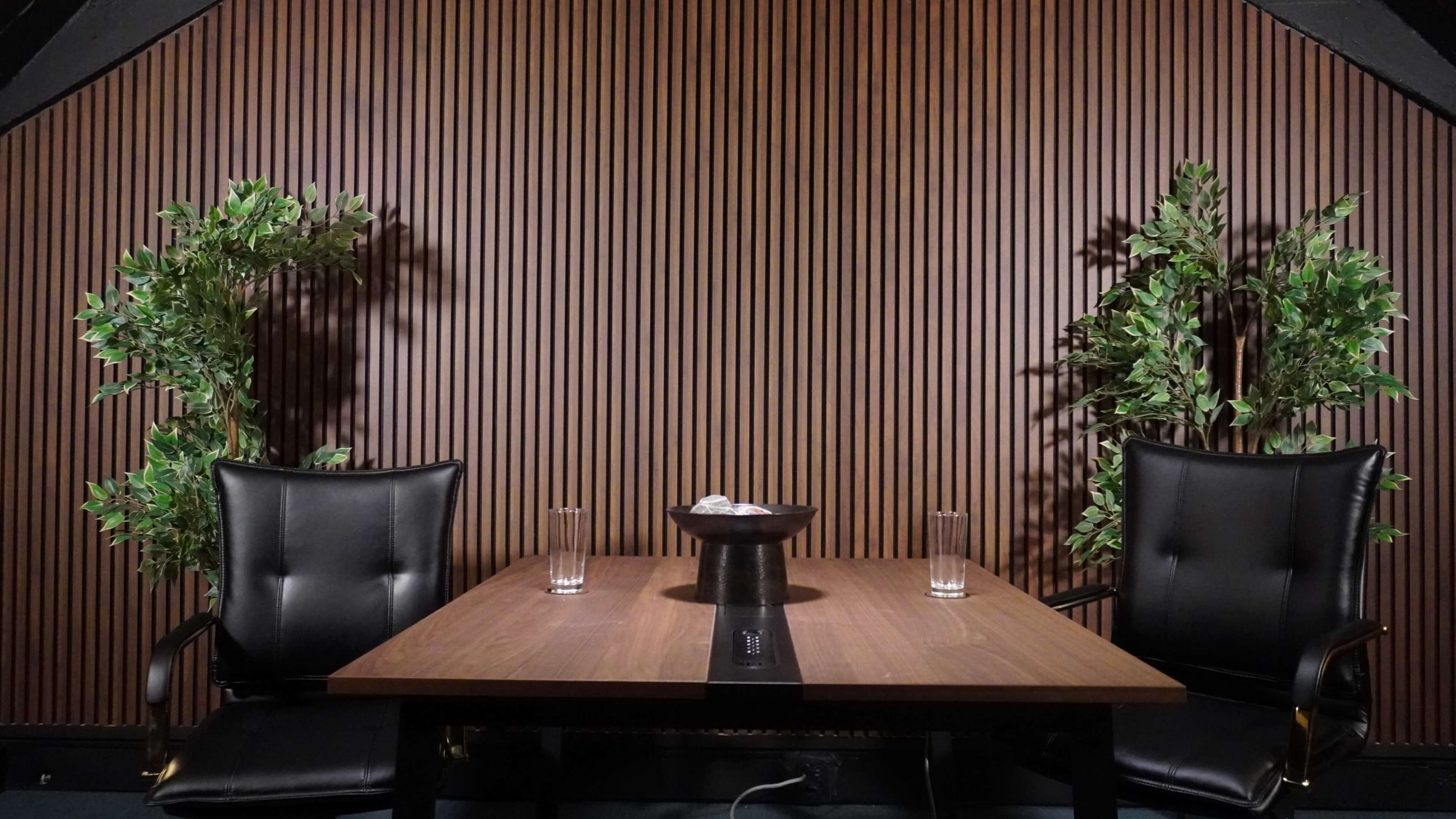 The image features a meeting room with a wooden table, two black chairs, and potted plants flanking the sides, set against a slatted wooden wall.
