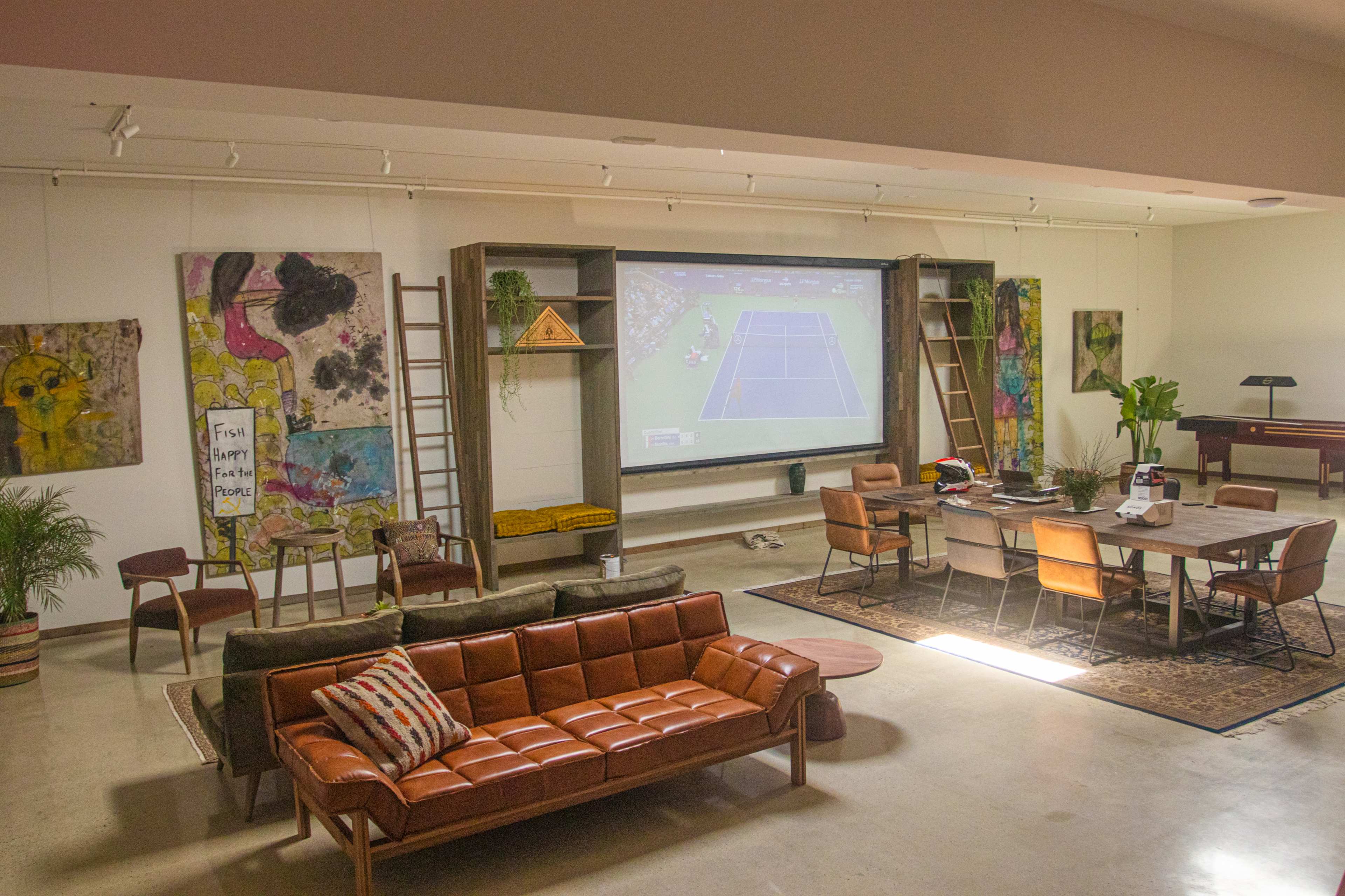 The image shows a spacious room with a large screen displaying a tennis match, surrounded by modern furniture, artwork on the walls, and a dining table set for a meal.