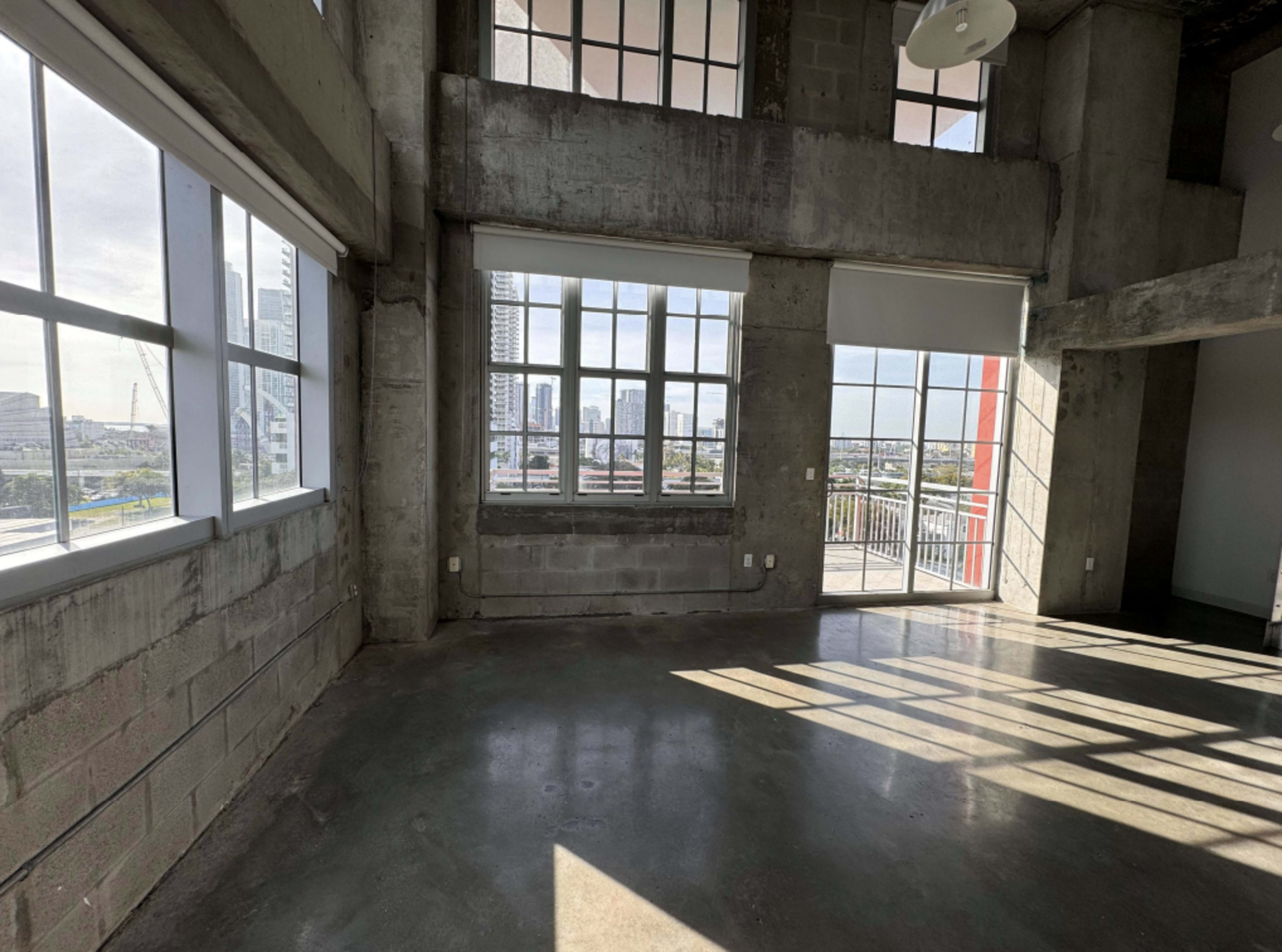 The image depicts a spacious, industrial-style room with large windows allowing natural light to fill the space and a view of a cityscape.
