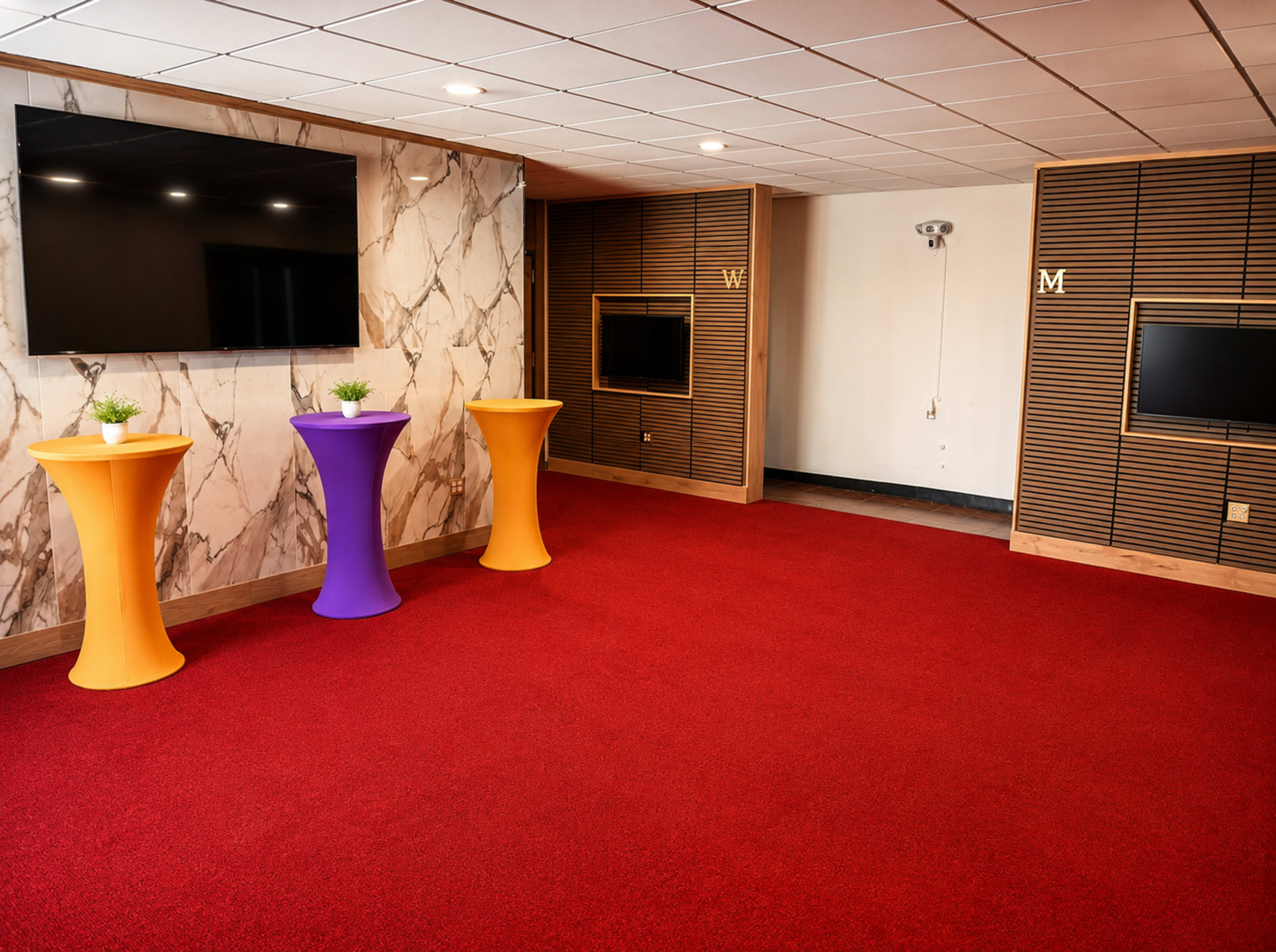 The image shows a brightly lit room featuring red carpeting, two wall-mounted televisions, and three colorful cocktail tables in orange, purple, and yellow.