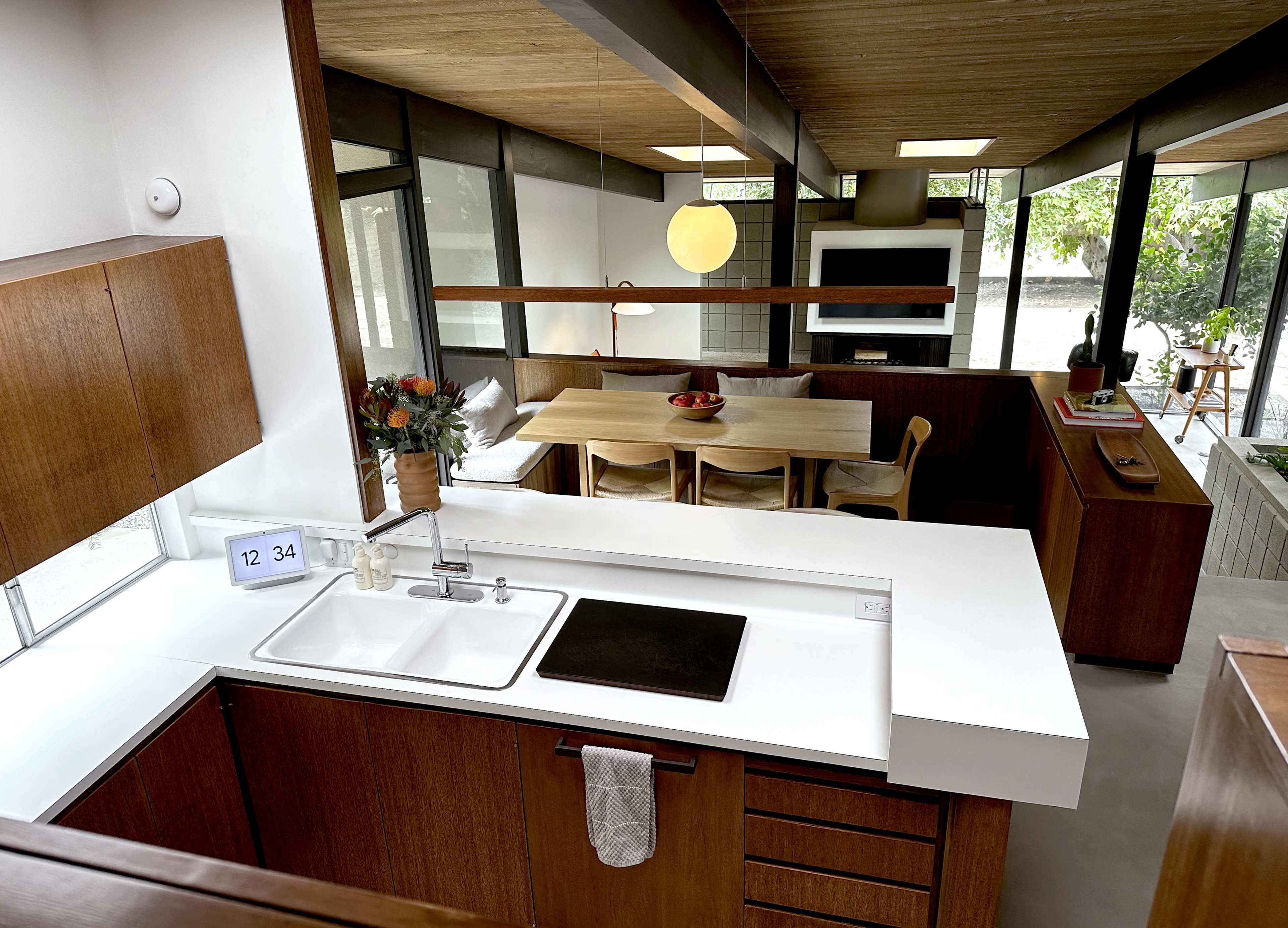 The image shows a modern kitchen with wooden cabinetry and a white countertop, adjacent to a dining area featuring a wooden table and chairs.