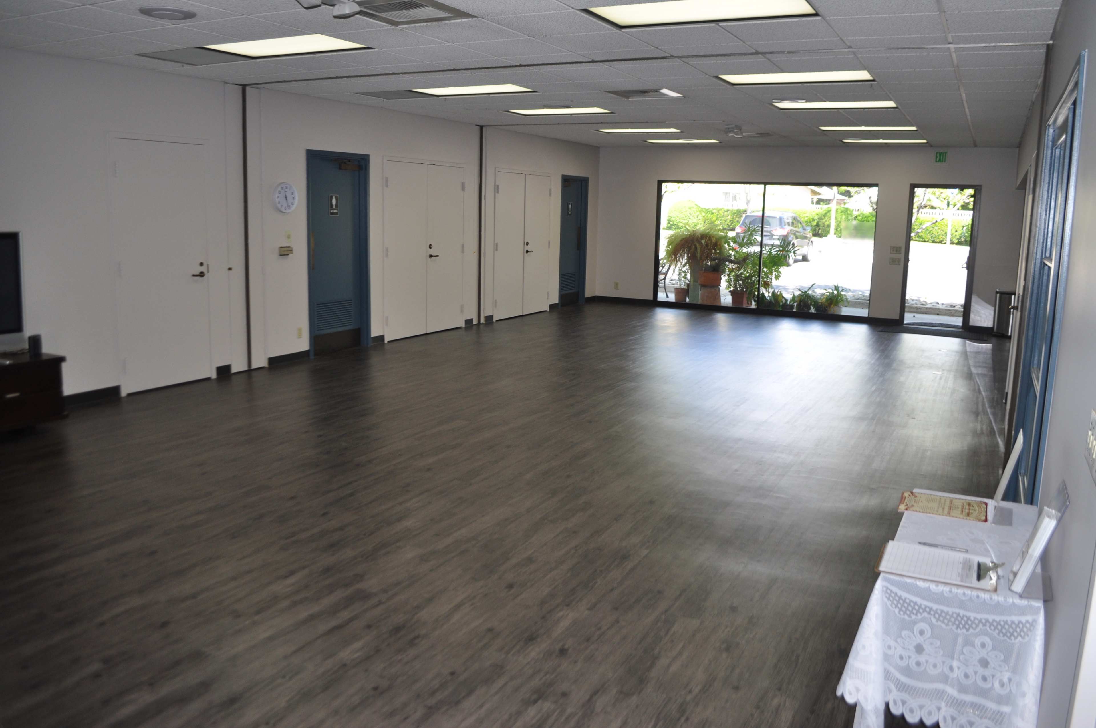 A large, empty indoor space with light-colored walls and a wooden floor, featuring several closed doors and a window that leads to an outdoor area.