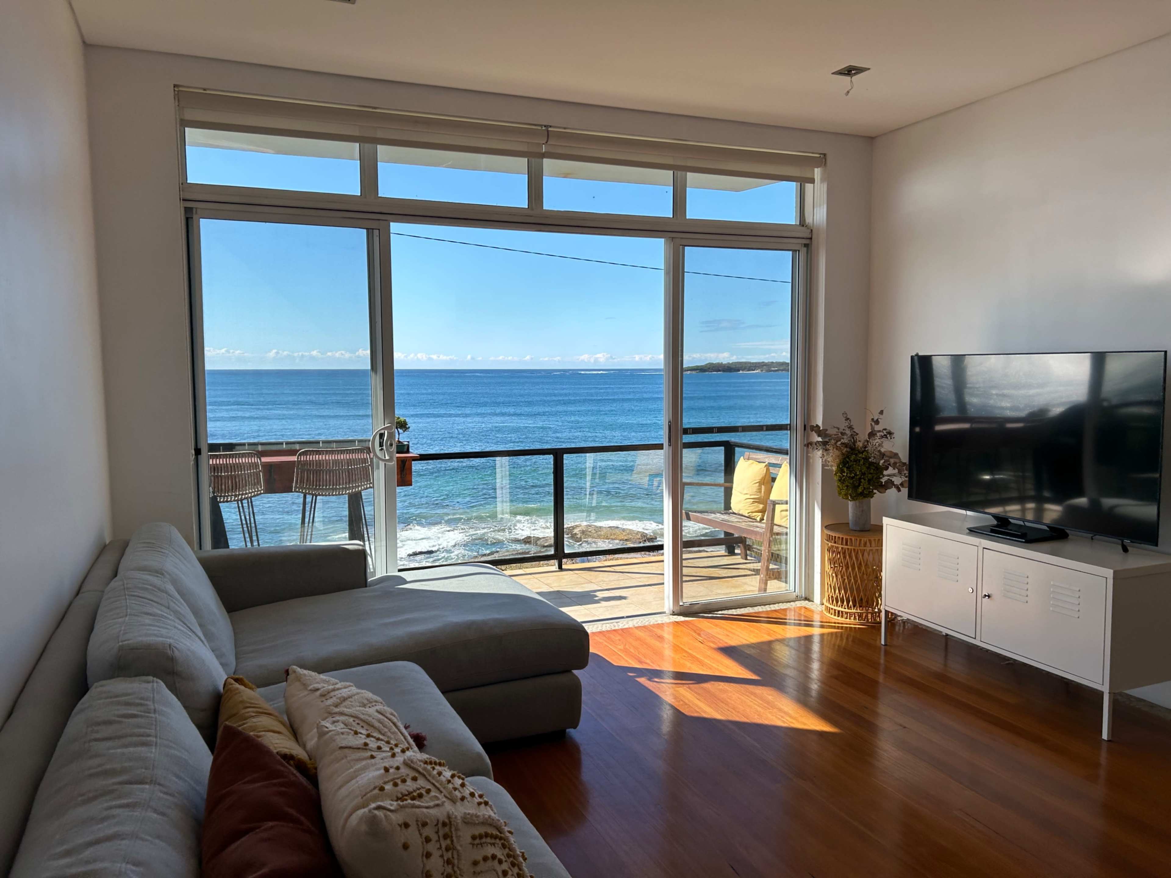A bright living room features a large window overlooking the ocean and a modern couch facing a television.