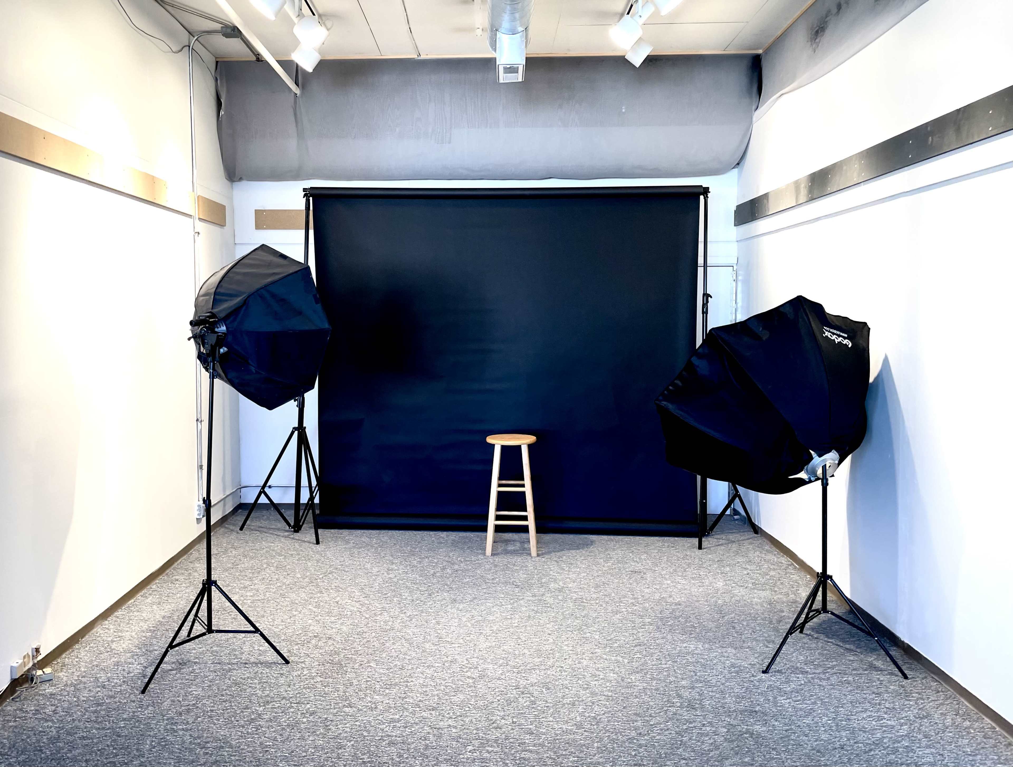 The image shows a photography studio featuring a black backdrop, a wooden stool in the center, and two softbox lights positioned on either side.