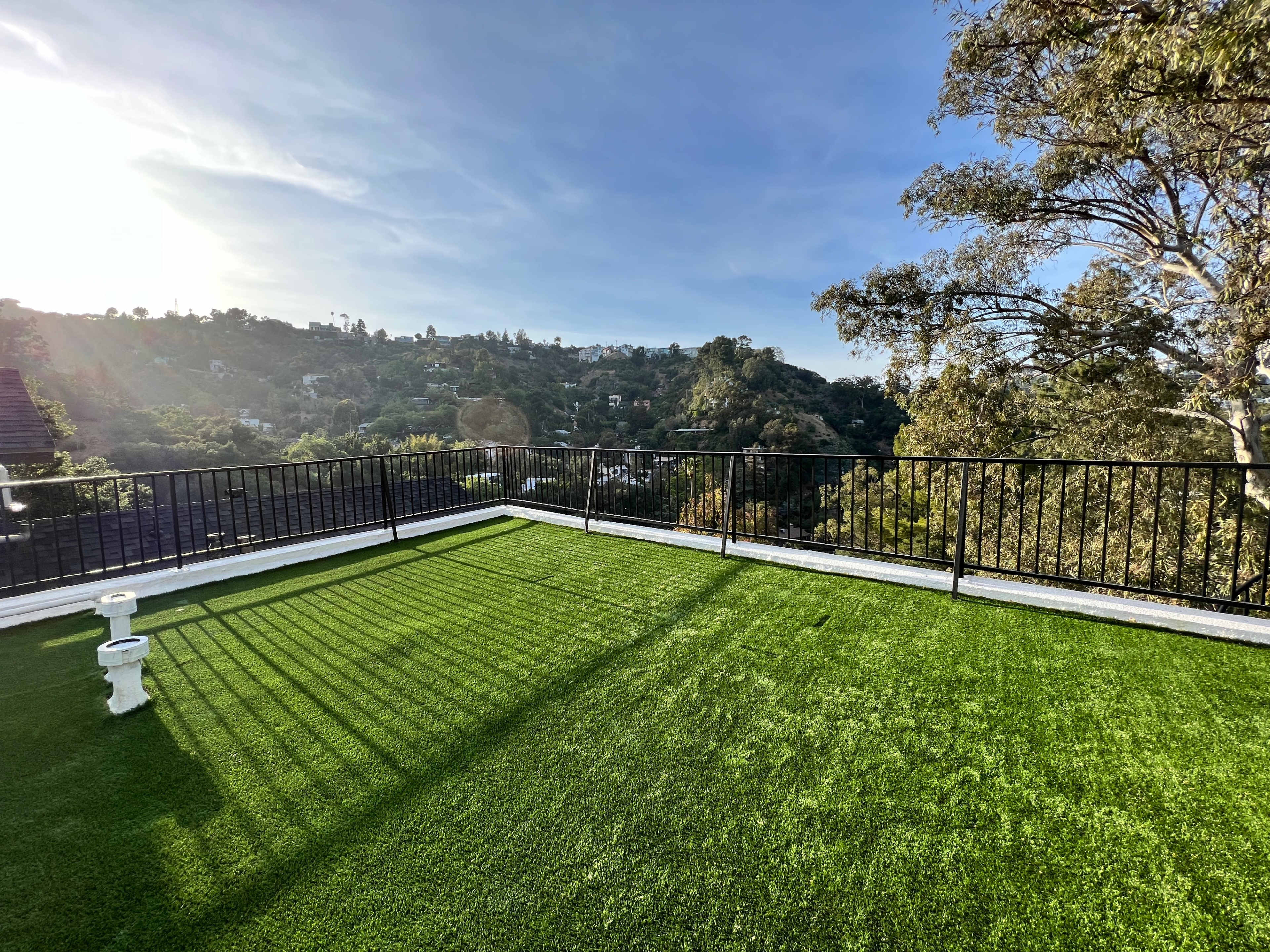A rooftop terrace with artificial grass overlooks a green hillside and trees under a clear sky.