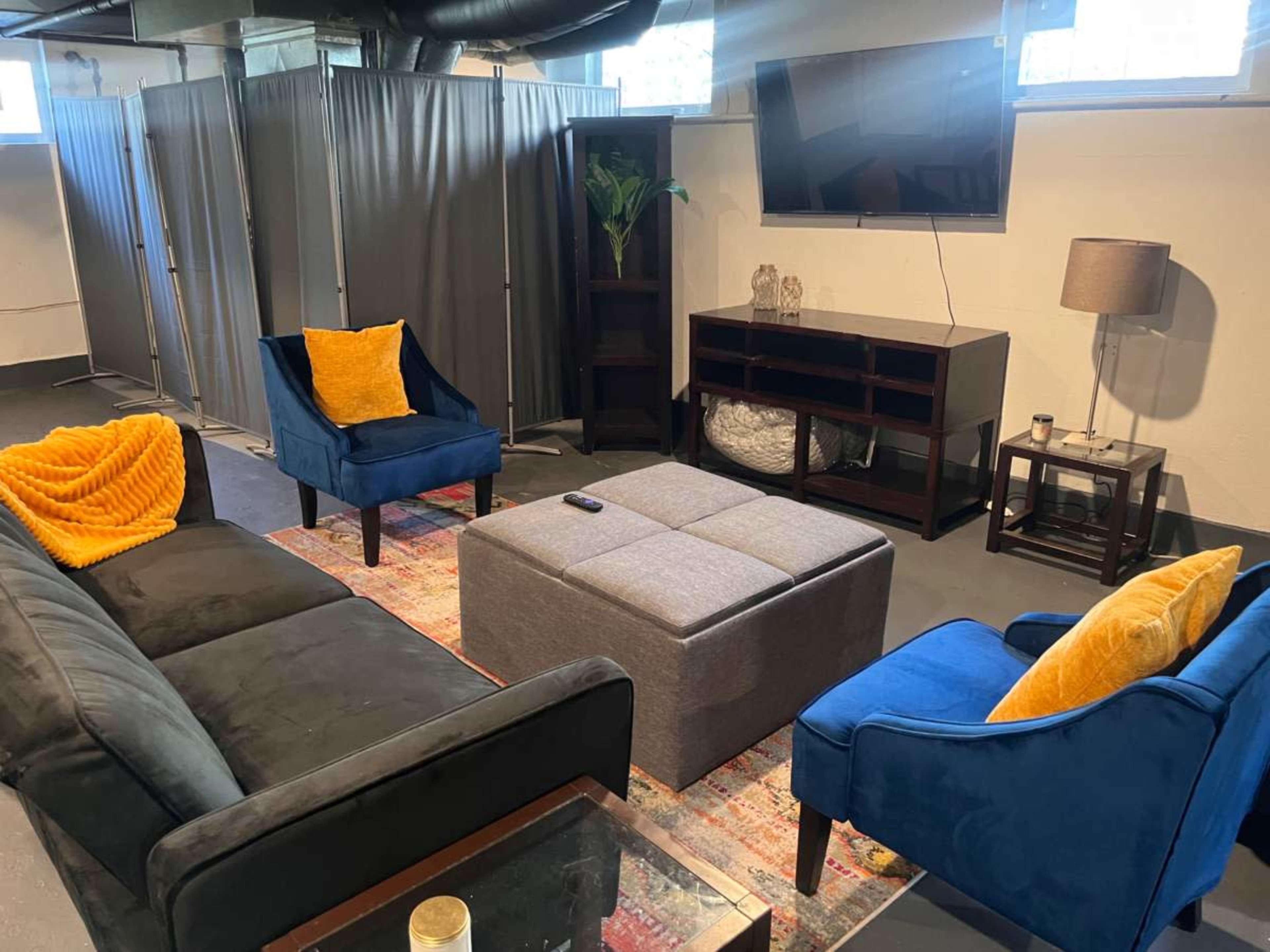 The image shows a modern living room setup with a dark sectional sofa, two blue armchairs, a gray ottoman, and a television mounted on the wall, surrounded by gray dividers.