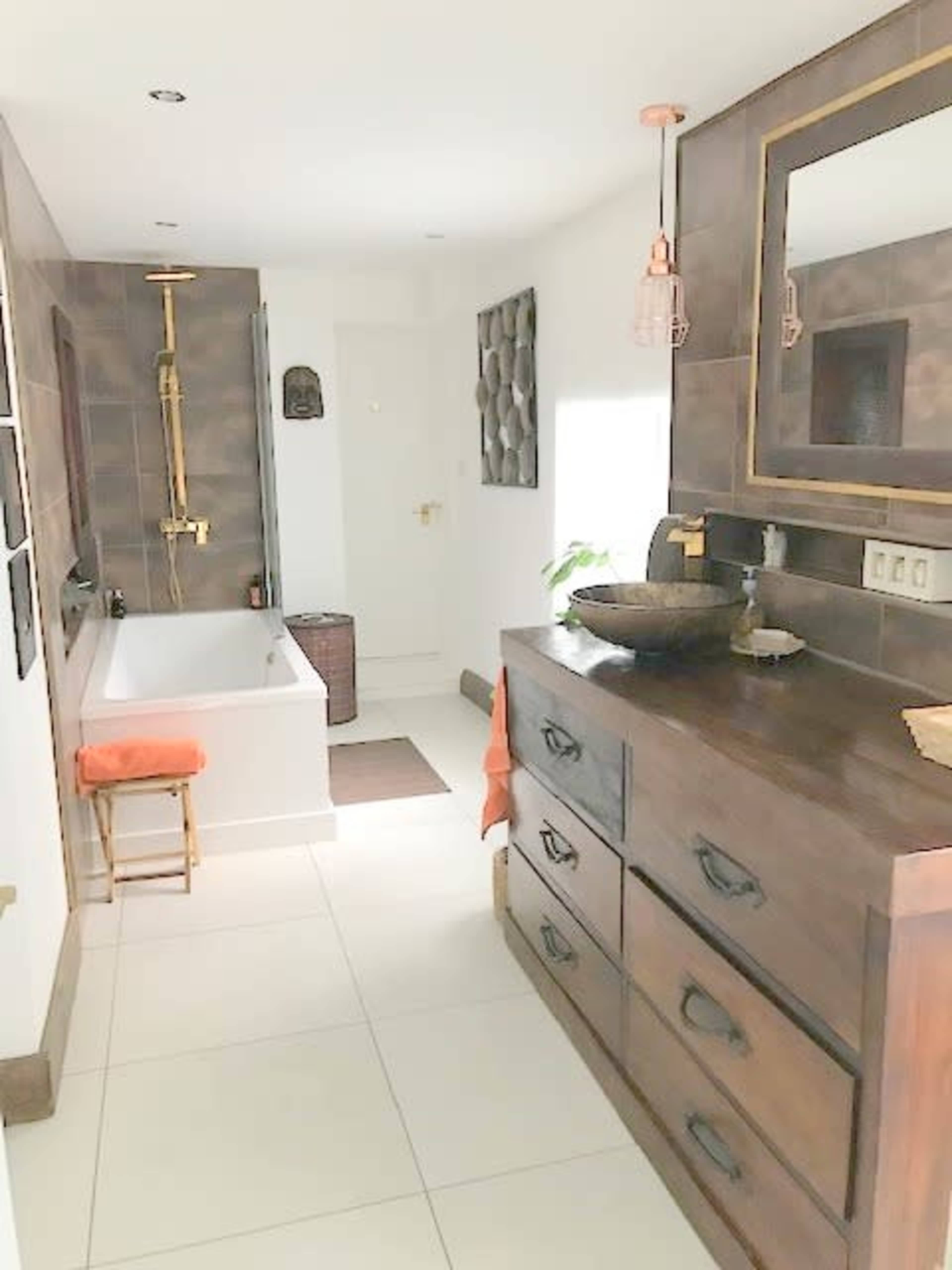 A modern bathroom featuring a bathtub, a wooden dresser with decorative items, and a well-lit layout with neutral tiles and fixtures.