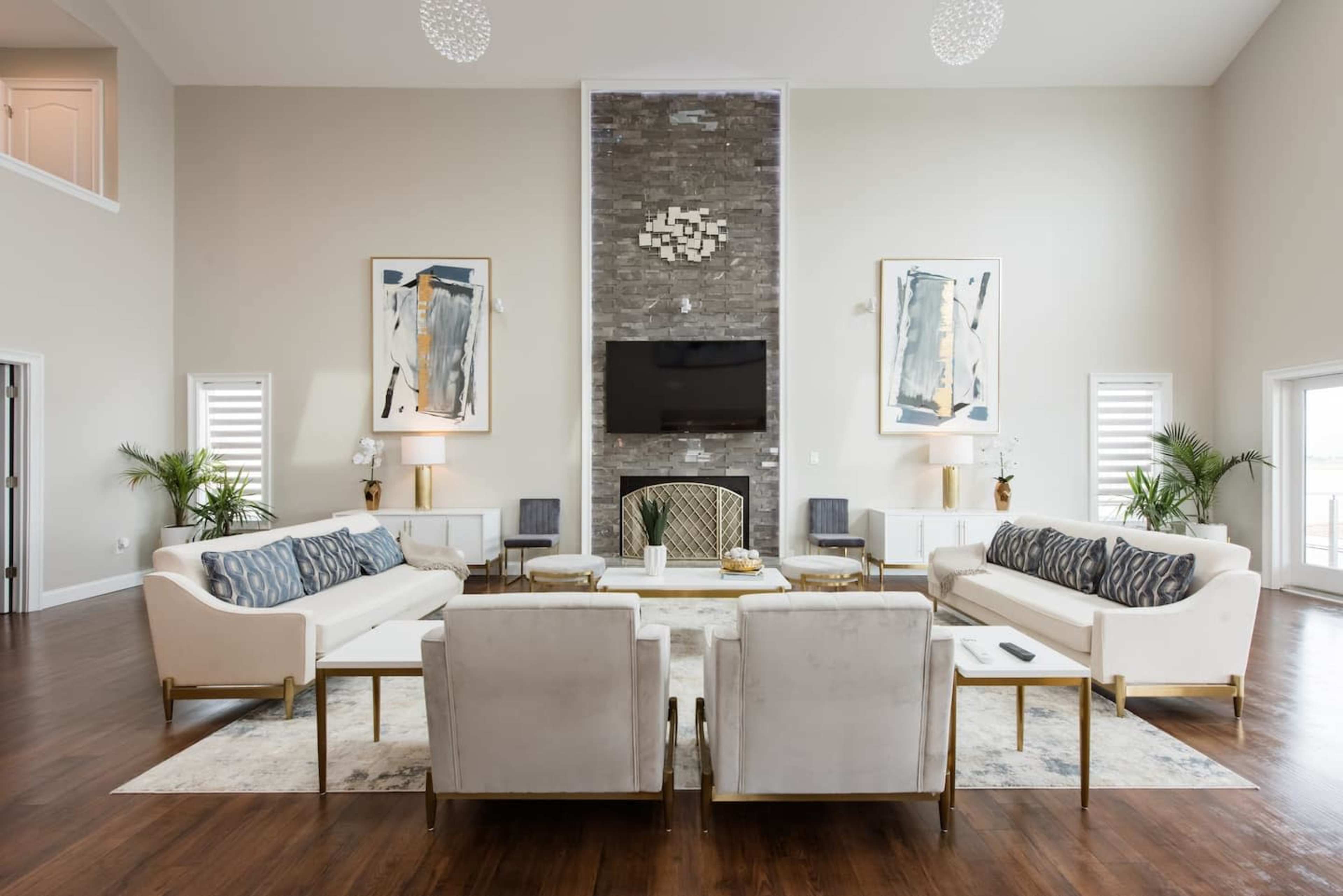 A spacious living room with two white sofas and two armchairs arranged around a central coffee table, a stone fireplace, and large artworks on the walls.