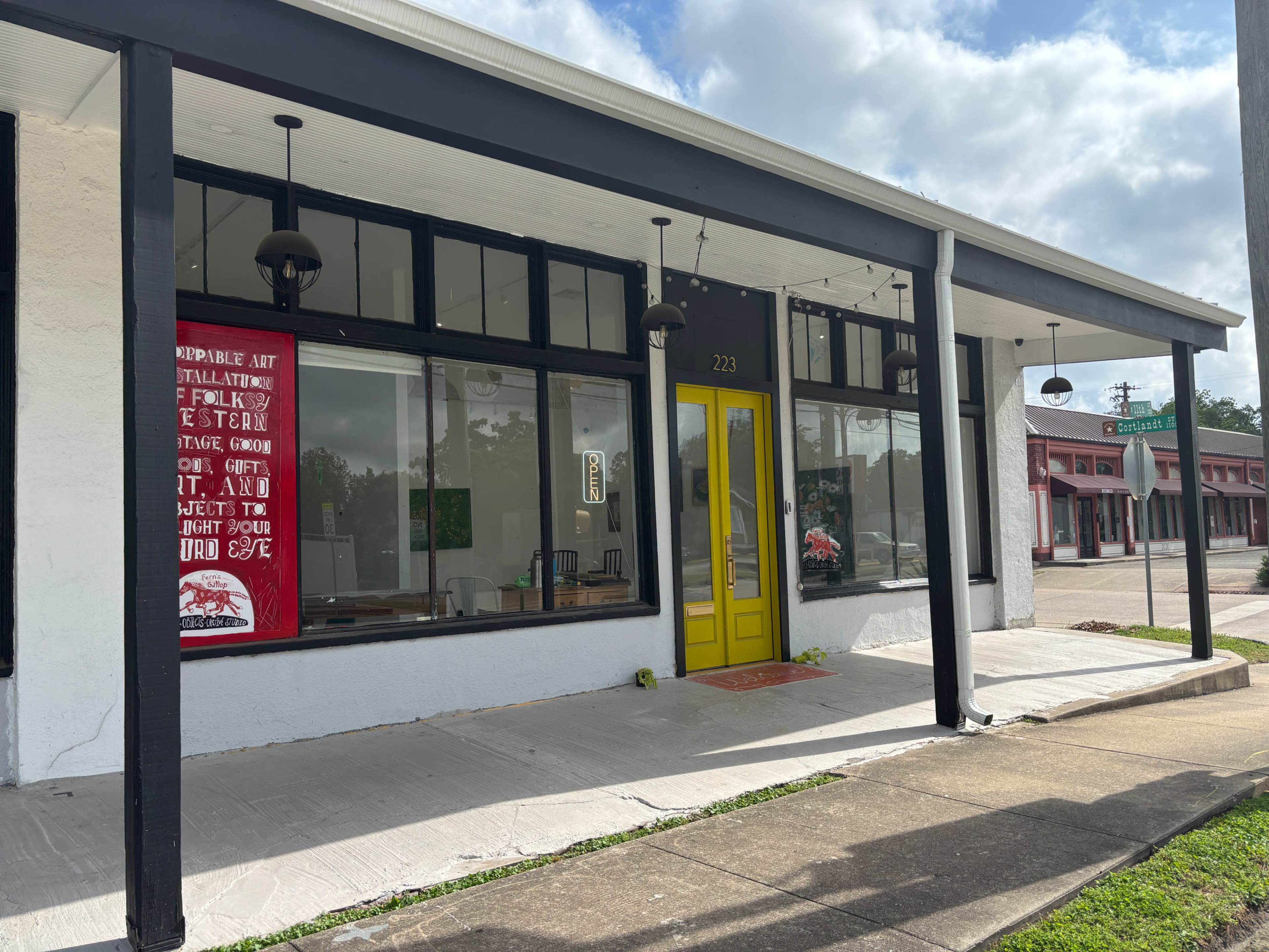 The image shows a storefront with a yellow door, large windows displaying art installations, and a banner hanging on the facade.