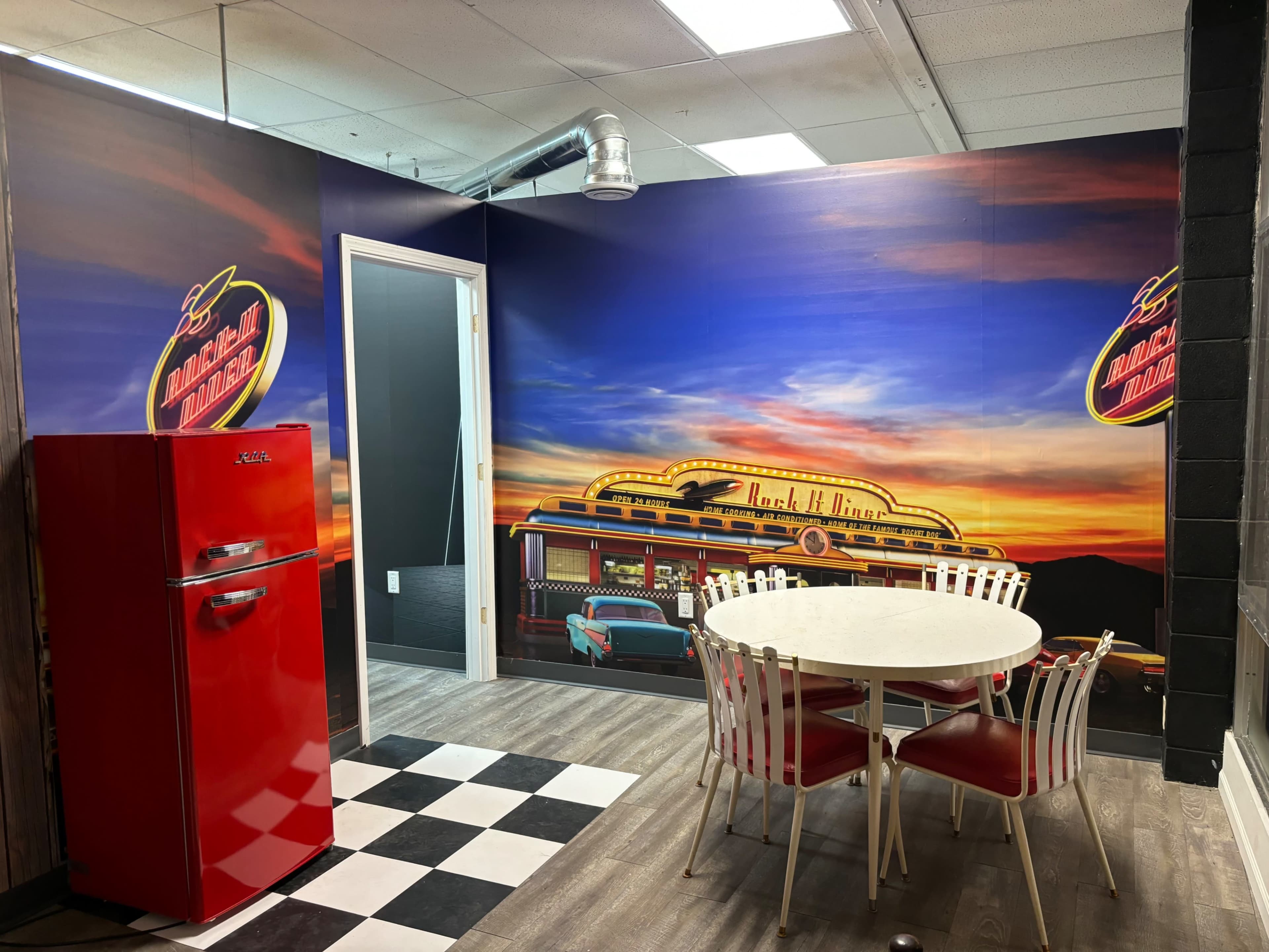 The image shows a retro-themed dining area with a red fridge, a round white table surrounded by white and red chairs, and a wall mural depicting a diner at sunset.
