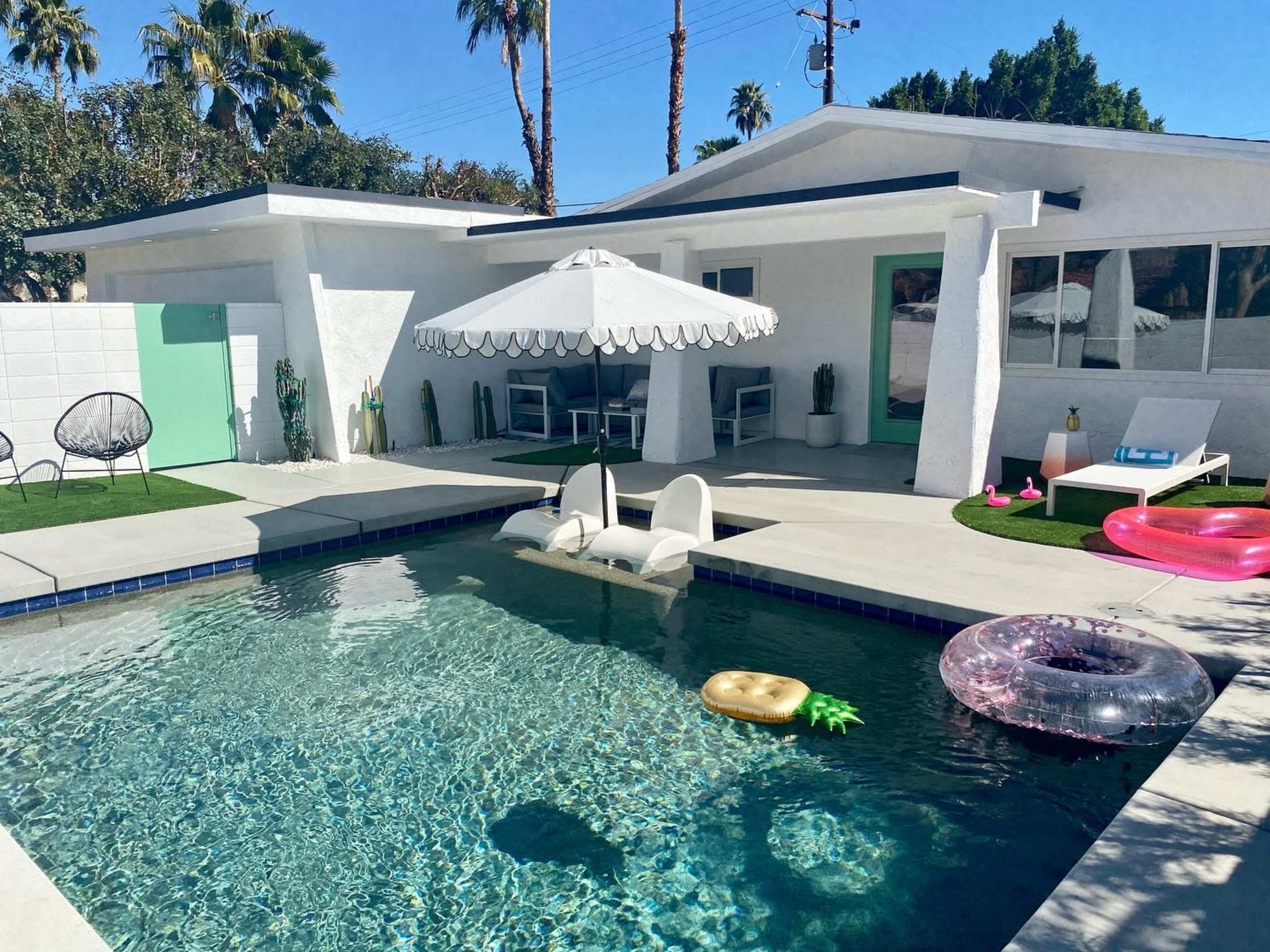 A modern backyard features a swimming pool surrounded by lounge chairs, an umbrella, and inflatable toys under clear blue skies.