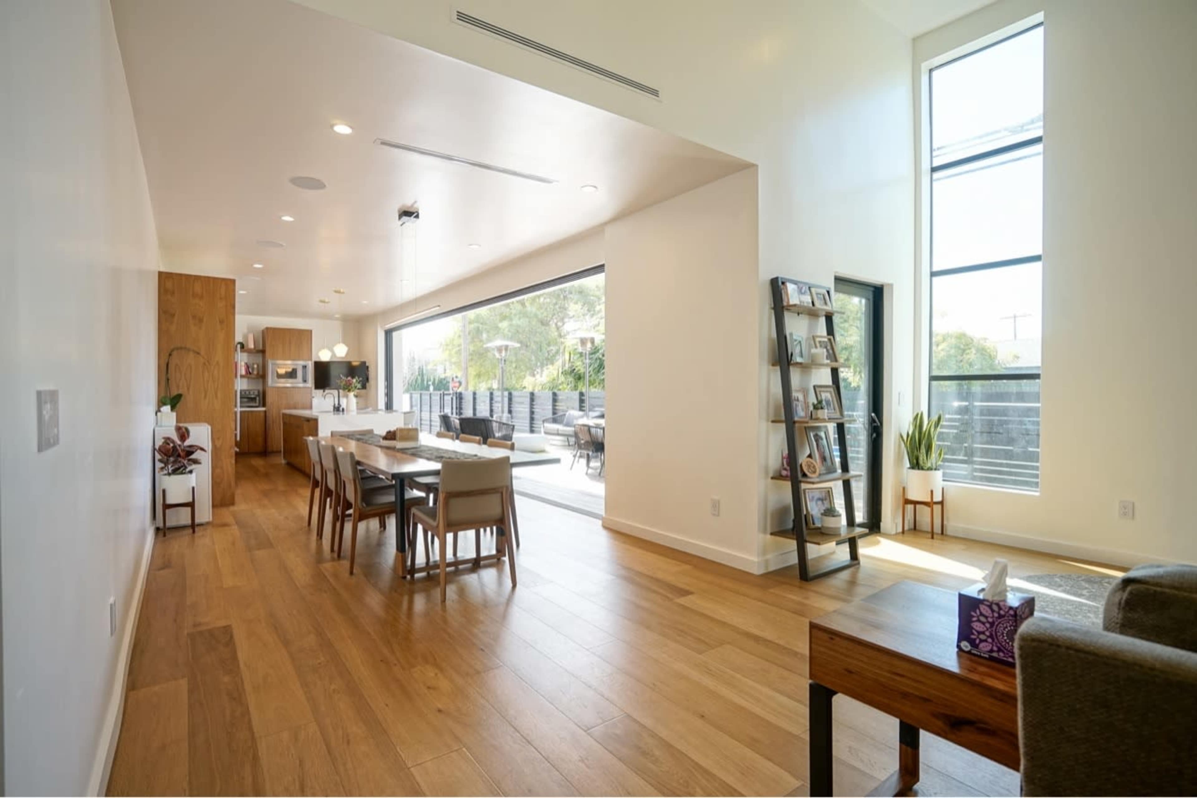The image shows a modern, open-concept living space with a dining area, kitchen, and large windows leading to an outdoor patio.
