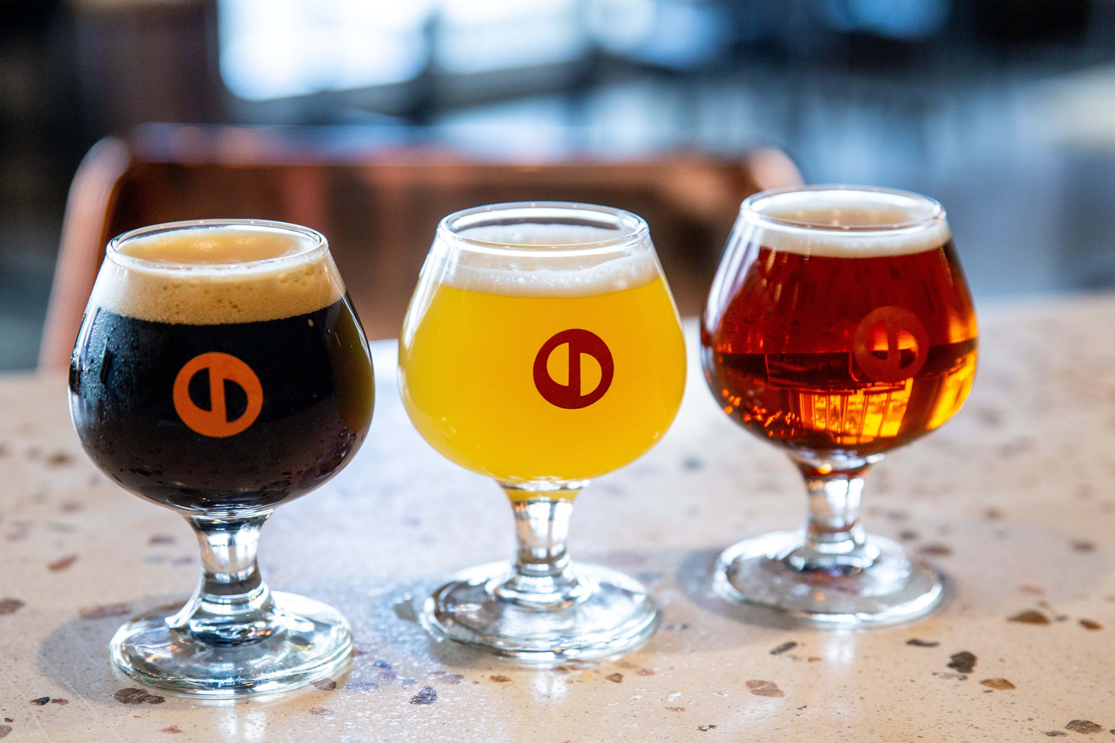 Three distinct glasses of beer are placed on a speckled countertop, featuring a dark brew, a golden lager, and a reddish amber beer, each with a circular logo on the side.
