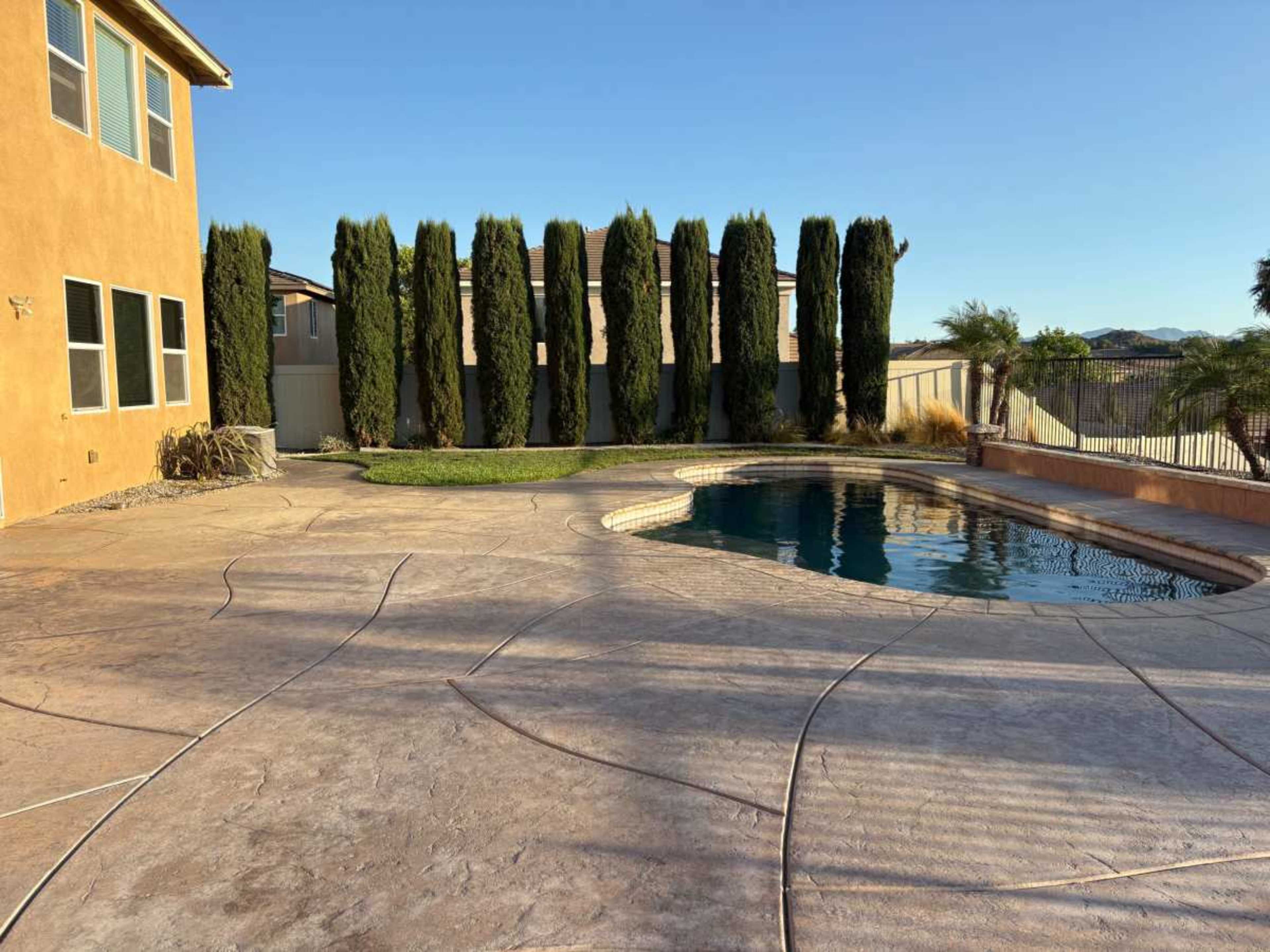 A backyard features a curved pool surrounded by stamped concrete and lined with tall, green cypress trees.