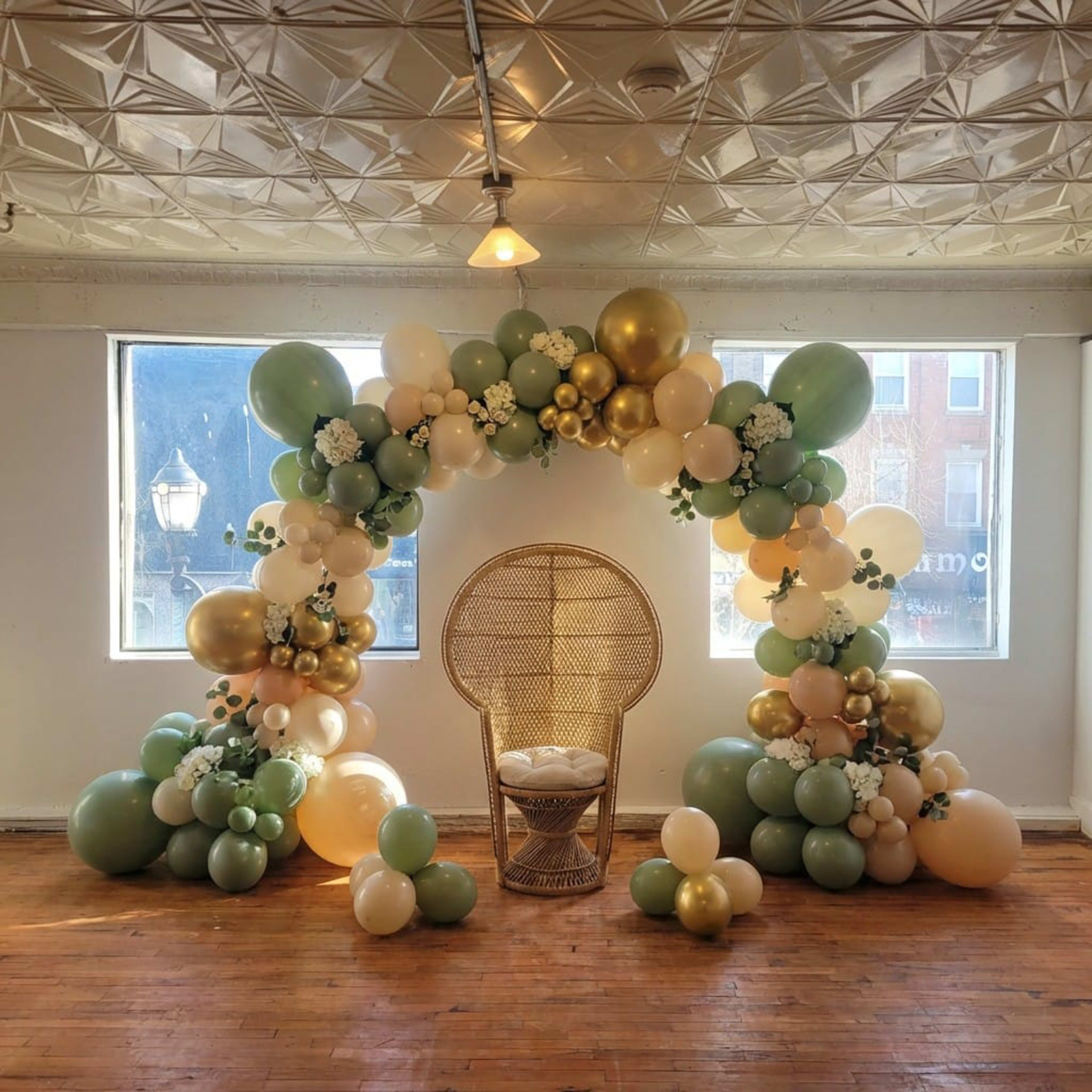 A decorative balloon arch in shades of green, gold, and cream frames a rattan chair against a backdrop of large windows.