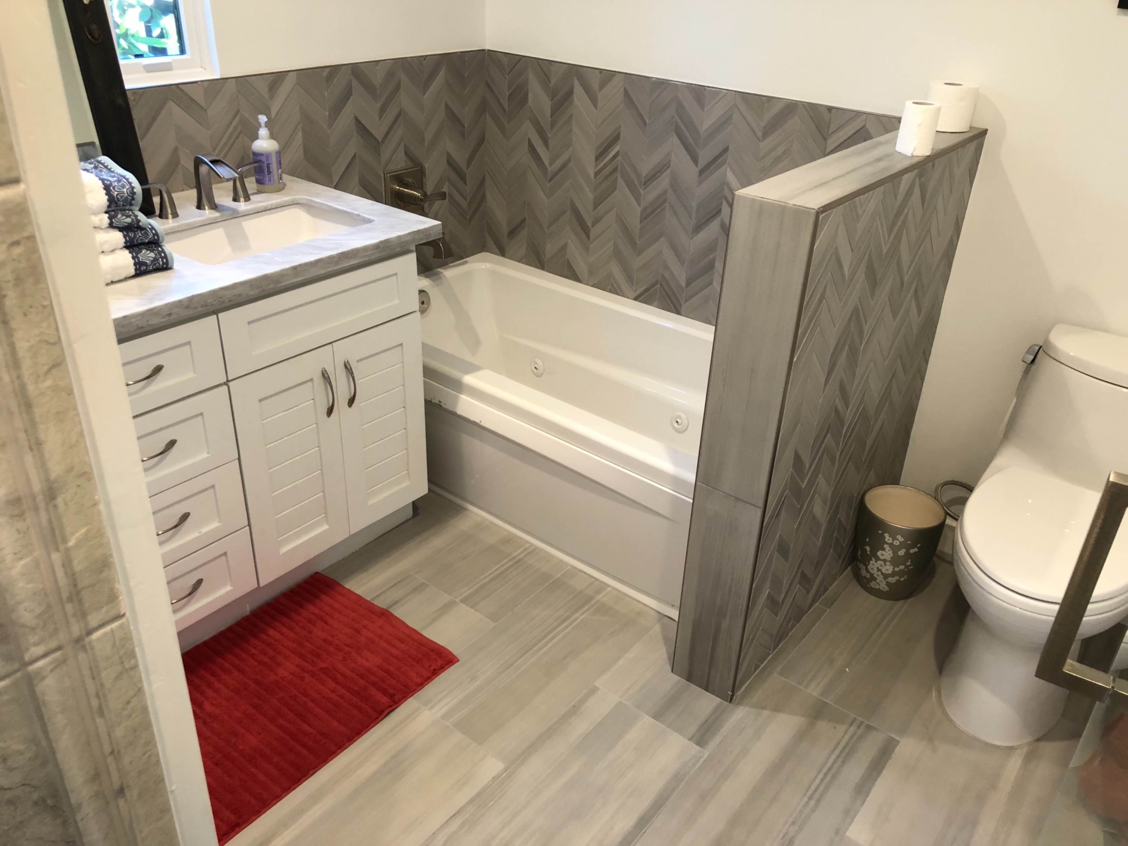 A modern bathroom features a tub with a sleek tile surround, a dual vanity with drawers, a toilet, and decorative towels on a red rug.