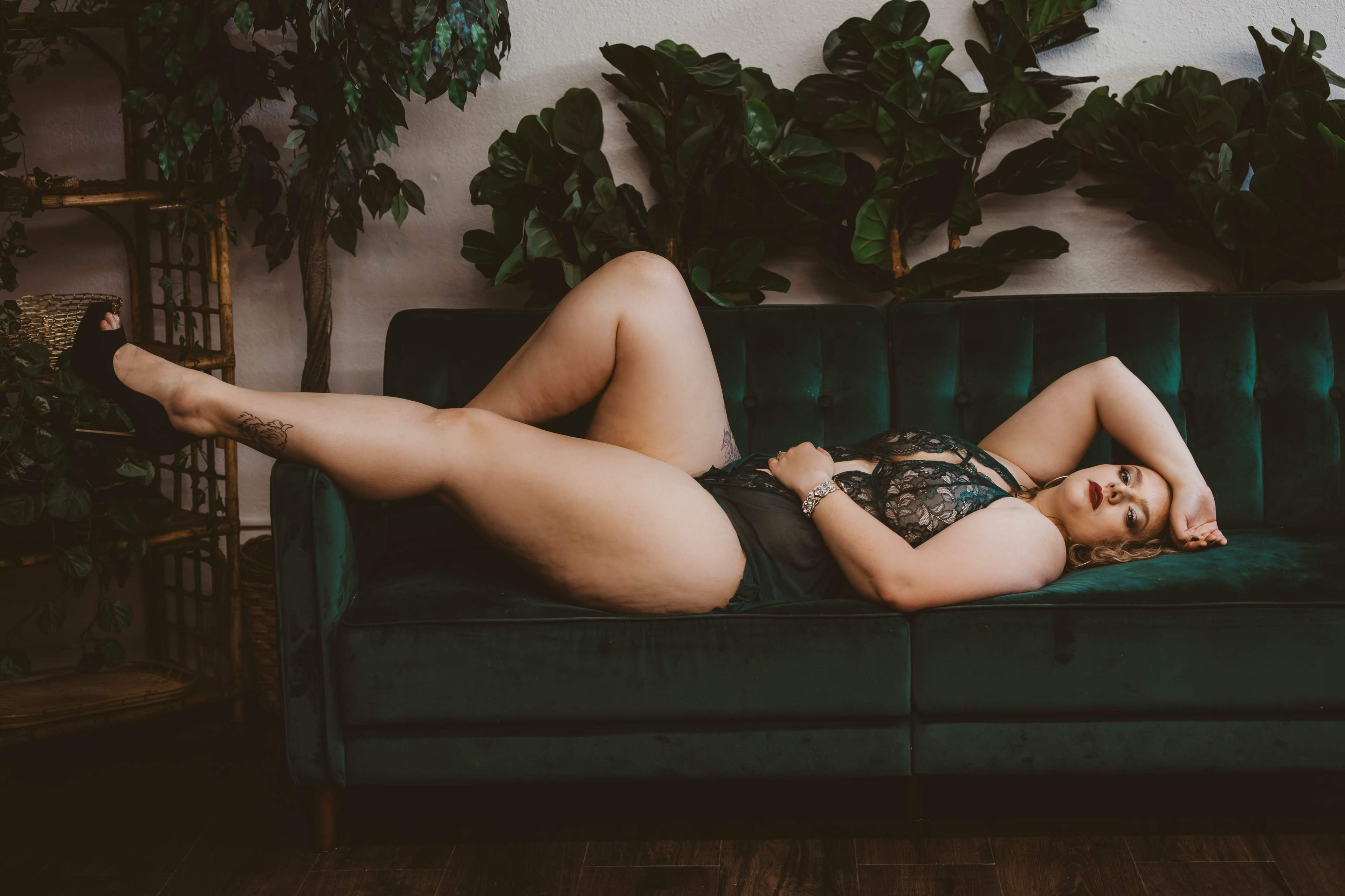 A person lies on a green velvet couch surrounded by large indoor plants.