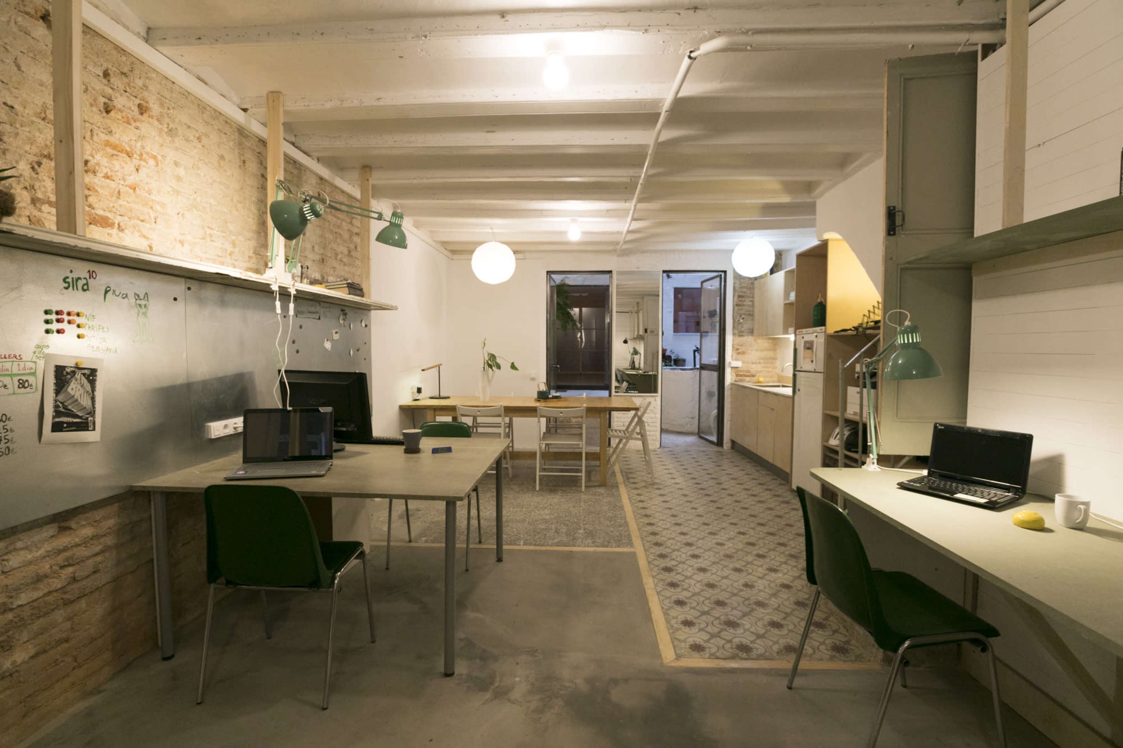 A spacious office with concrete floors, wooden beams, and a mix of desks, laptops, and kitchen areas, illuminated by overhead lights and green lamps.