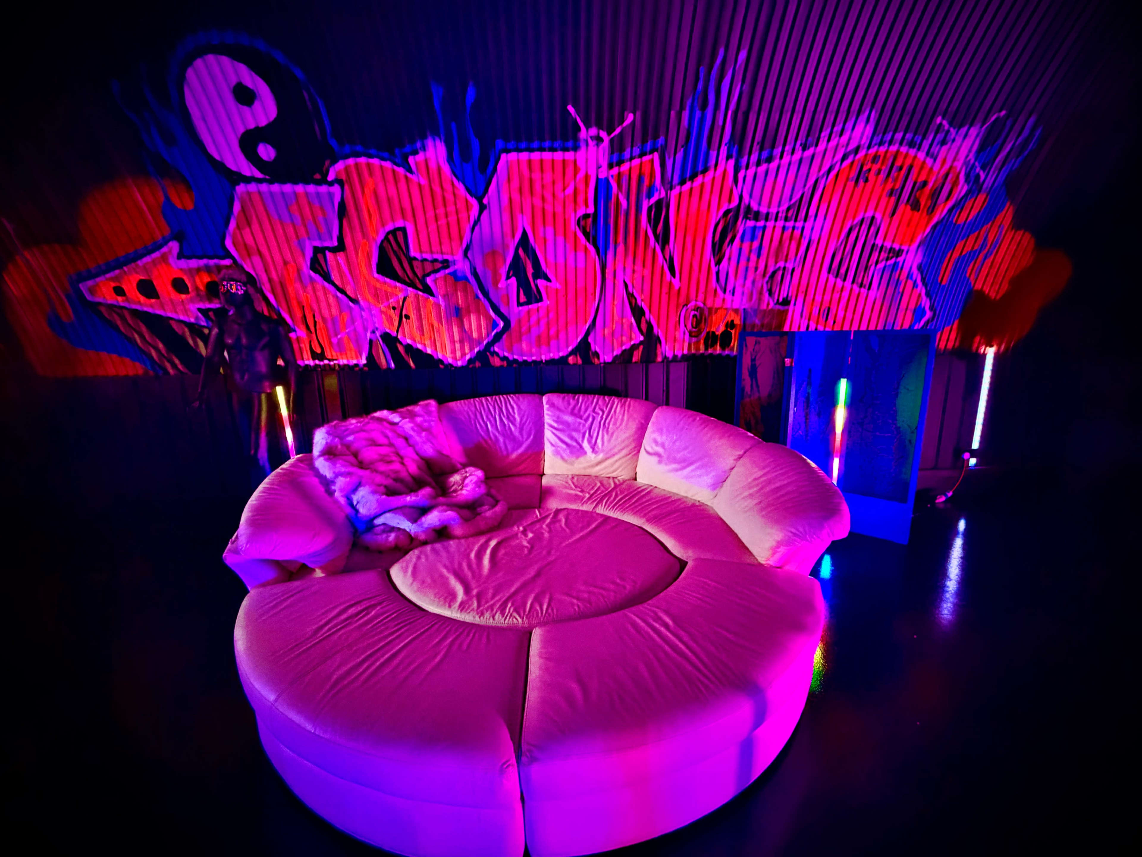 A circular, white sofa is positioned in front of a brightly colored graffiti wall featuring the word "GONE" and illuminated by colorful lights.