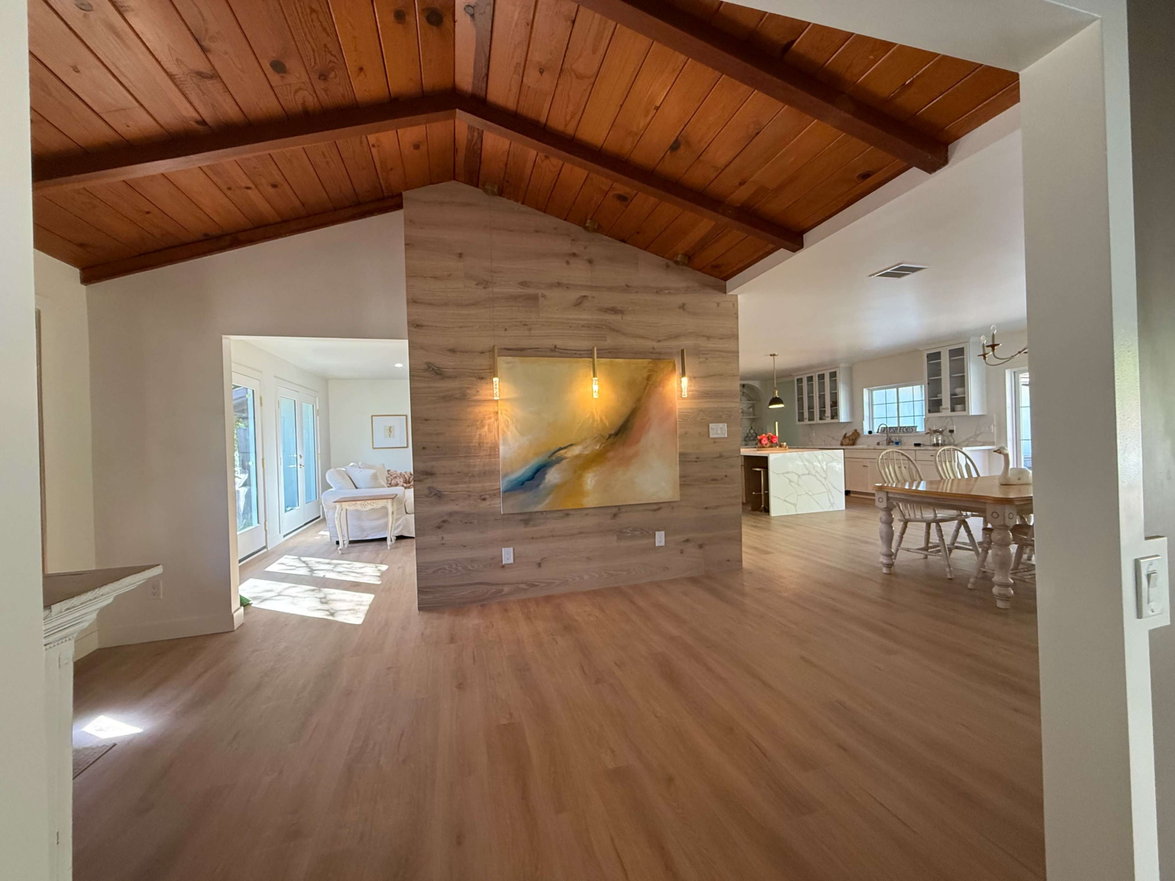 The image shows an interior space featuring a wood-beamed ceiling, a wooden accent wall with a painting, and an open layout connecting to a kitchen area in the background.
