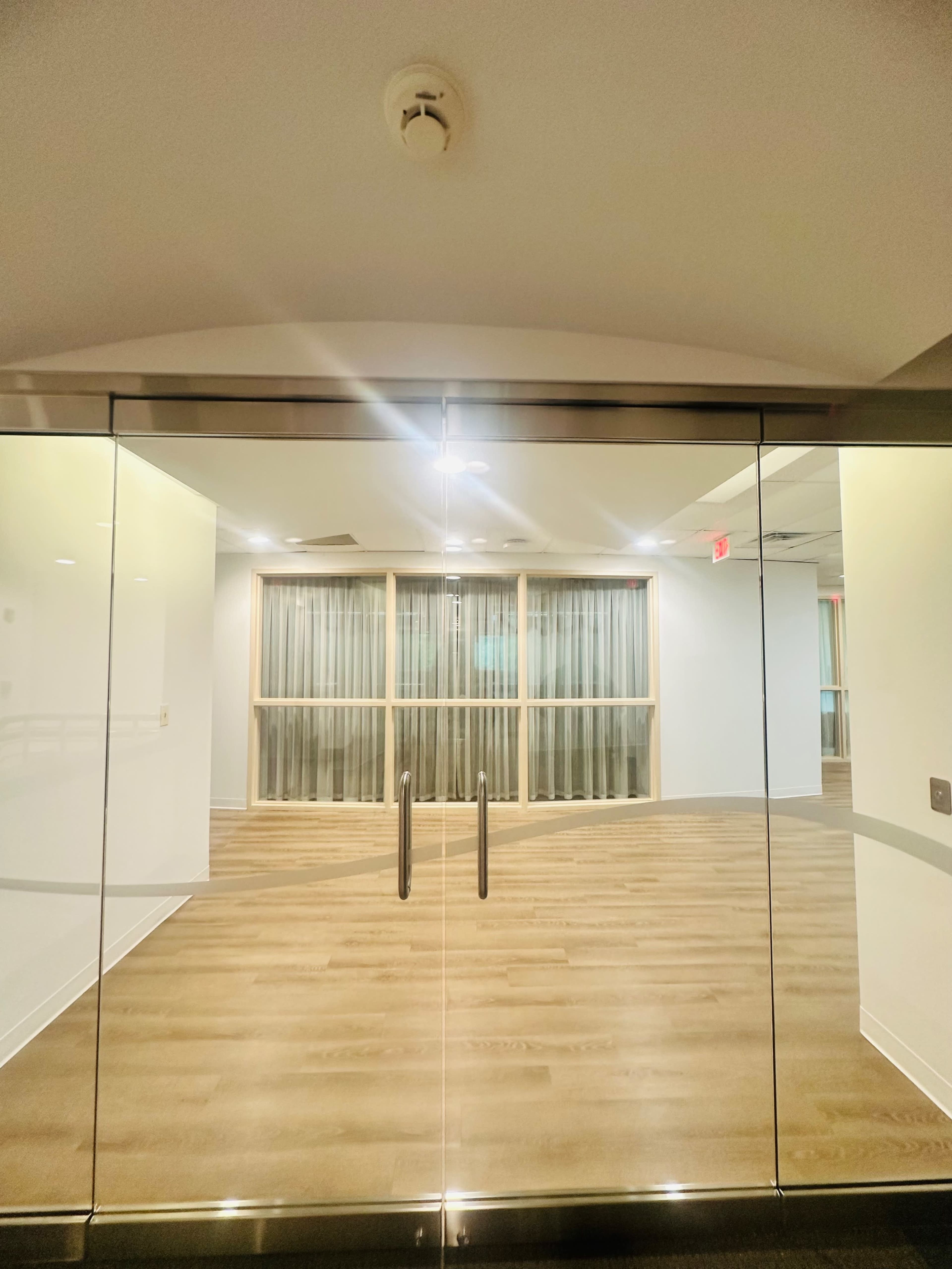 The image shows a glass entrance leading into a room with wooden flooring and large windows covered by sheer curtains.