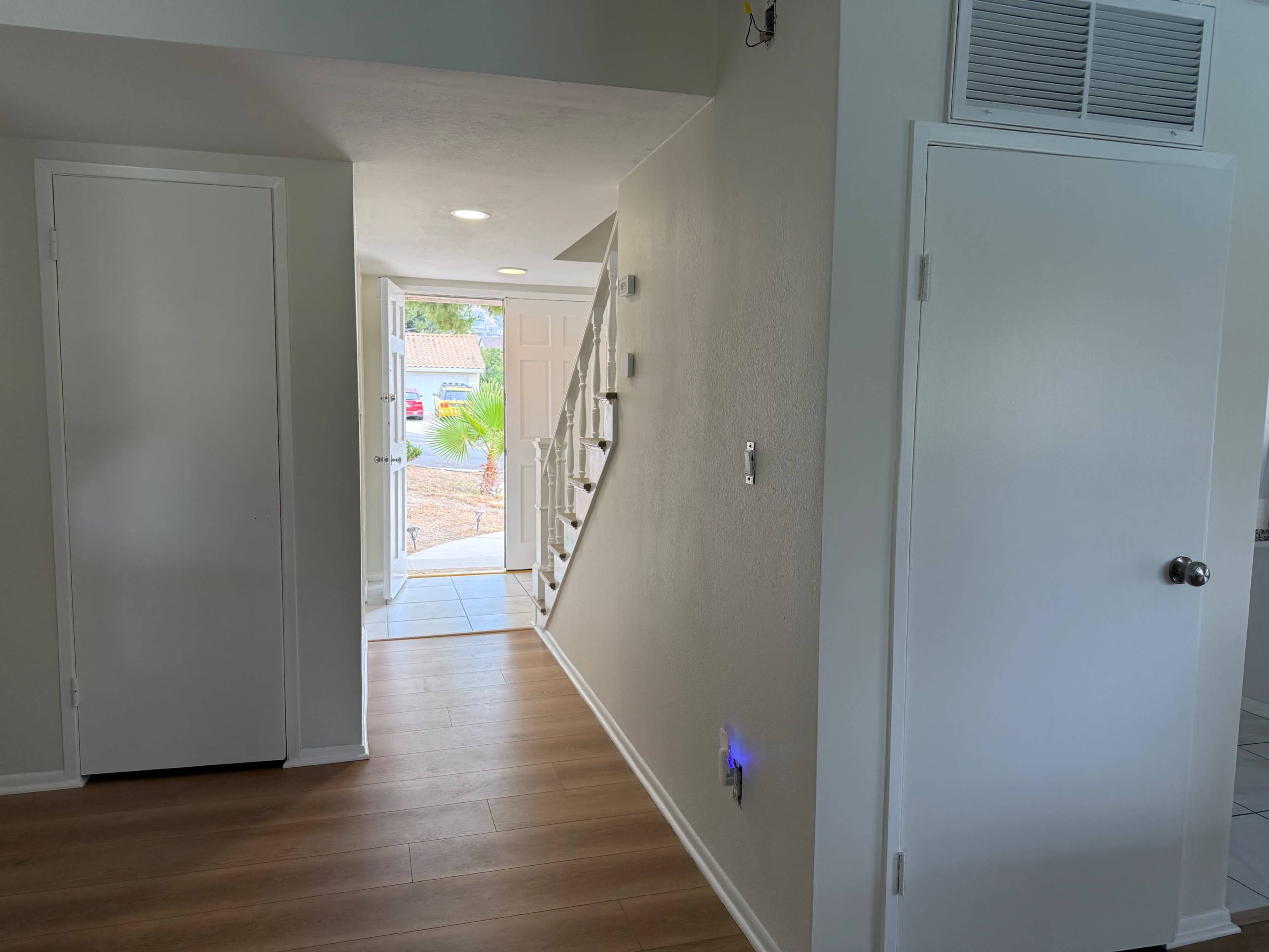 The image shows a hallway leading to a staircase, with a door open to an outside area, and light-colored walls and flooring.