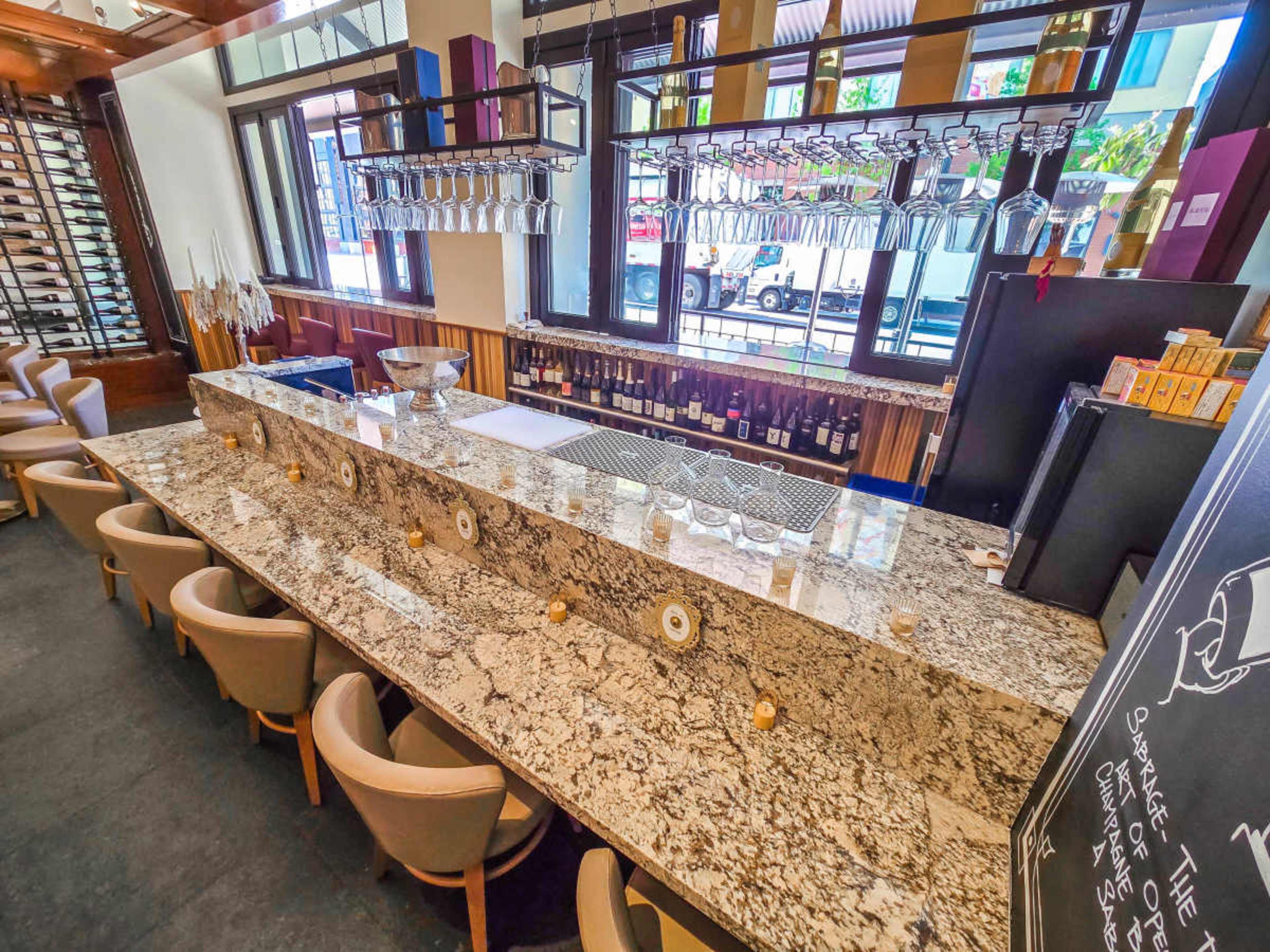 The image shows a bar area with a long granite countertop, neatly arranged with glasses hanging above and various bottles displayed behind.