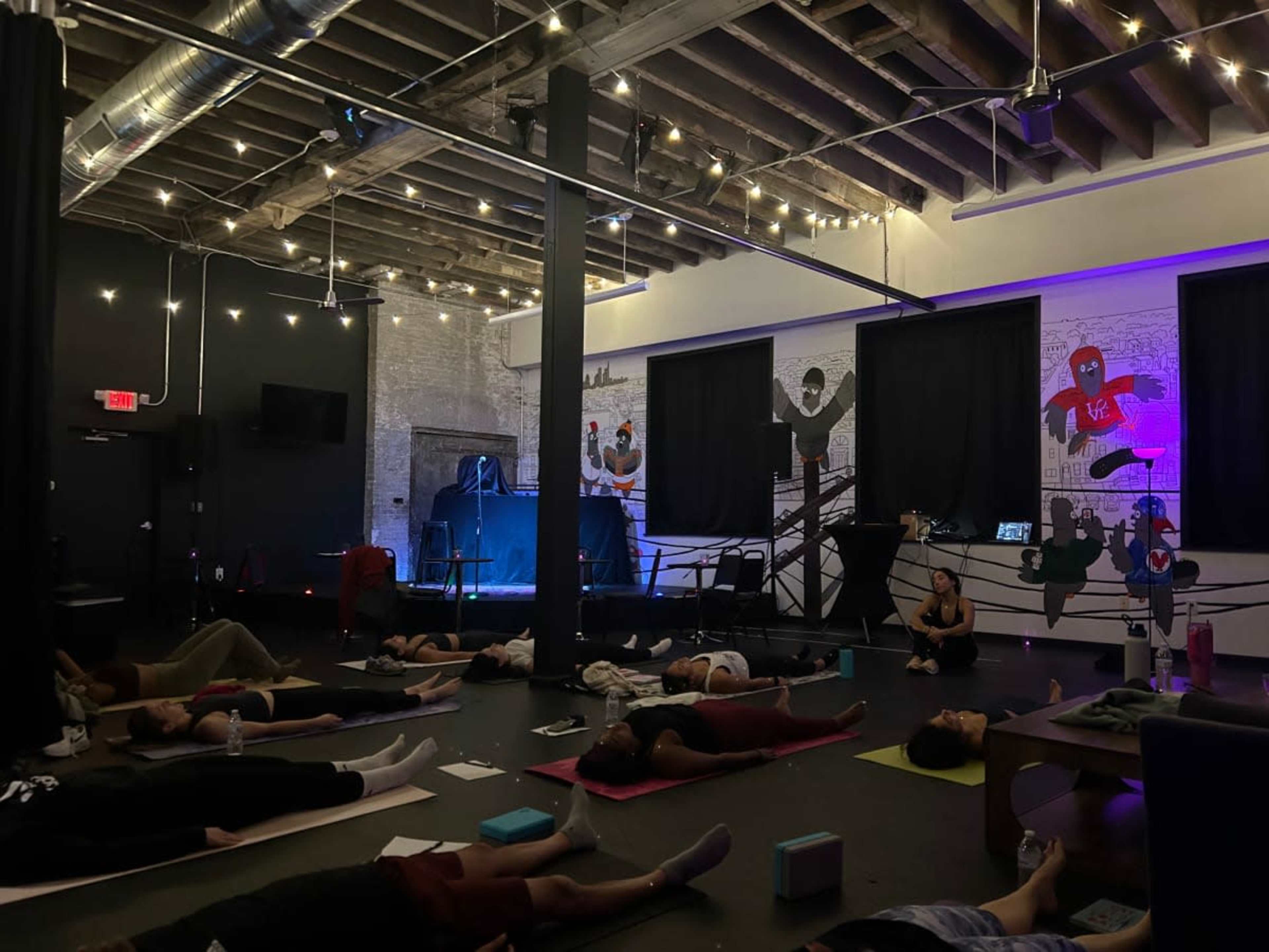 A group of people practice yoga on mats inside a dimly lit studio featuring exposed beams and string lights, while a mural adorns one wall.