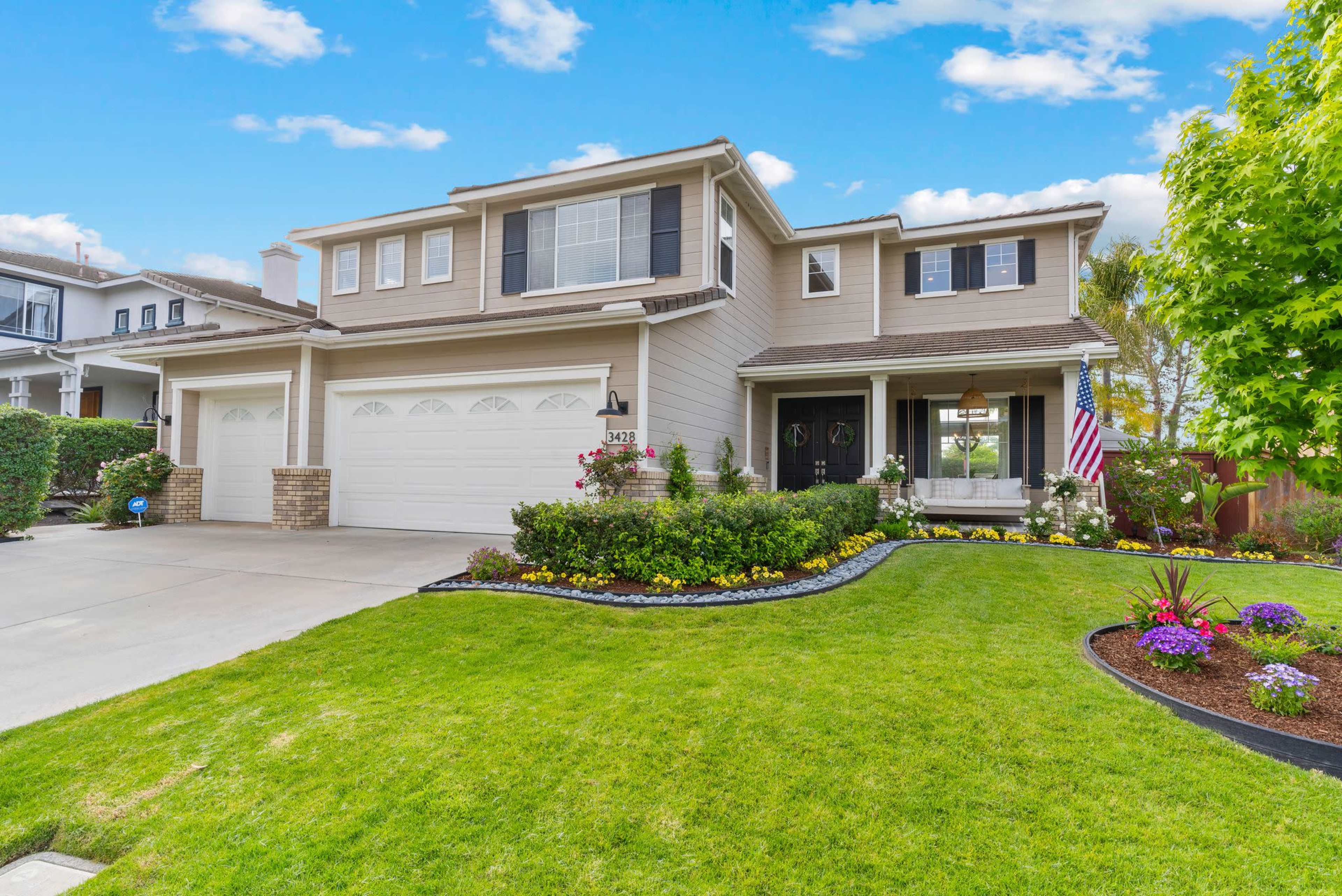 A two-story suburban house with a well-maintained lawn, colorful flowerbeds, and a paved driveway leading to a double garage.