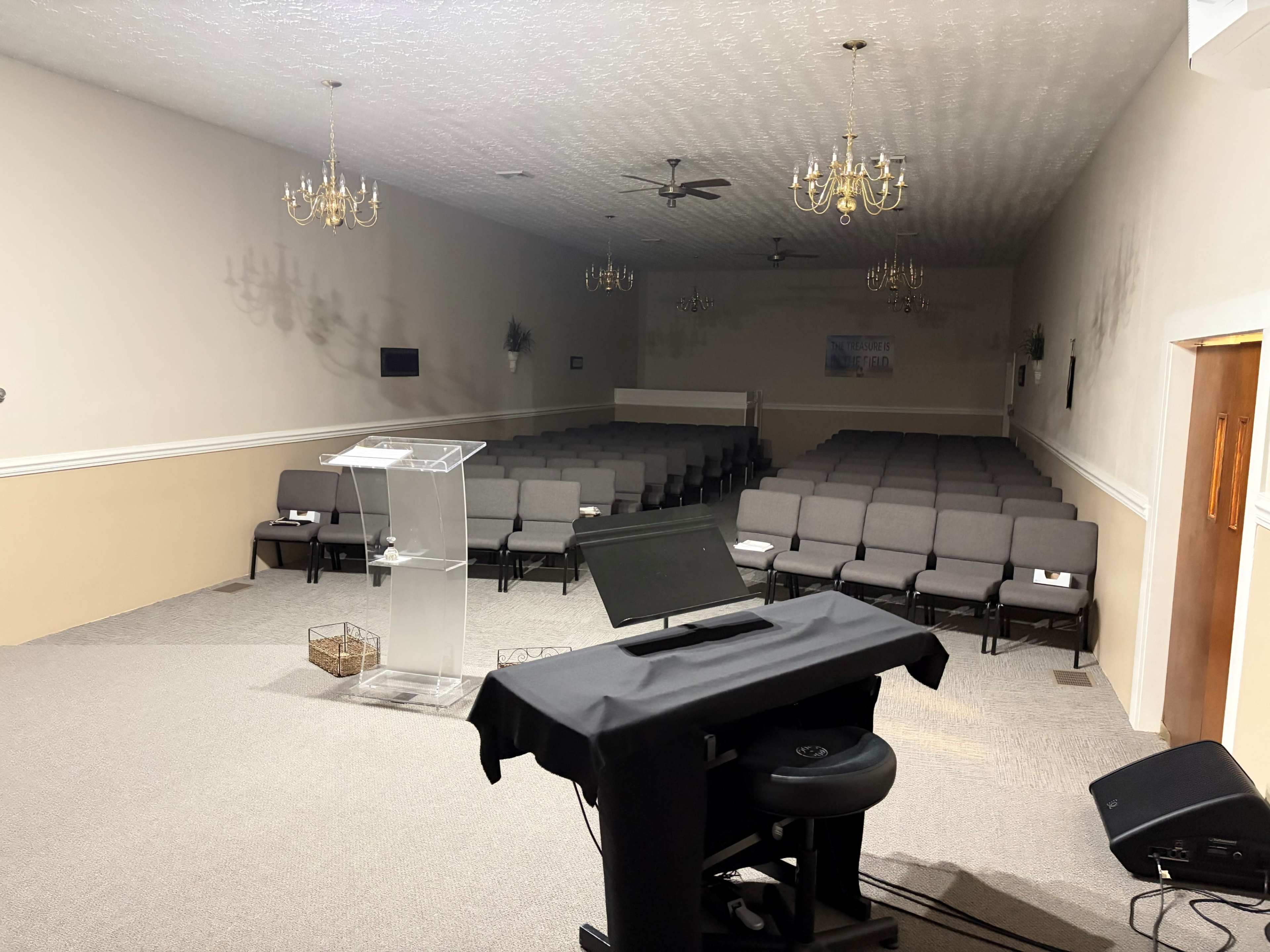 The image shows an empty church sanctuary with rows of gray chairs facing a clear podium and a sound setup in the foreground.