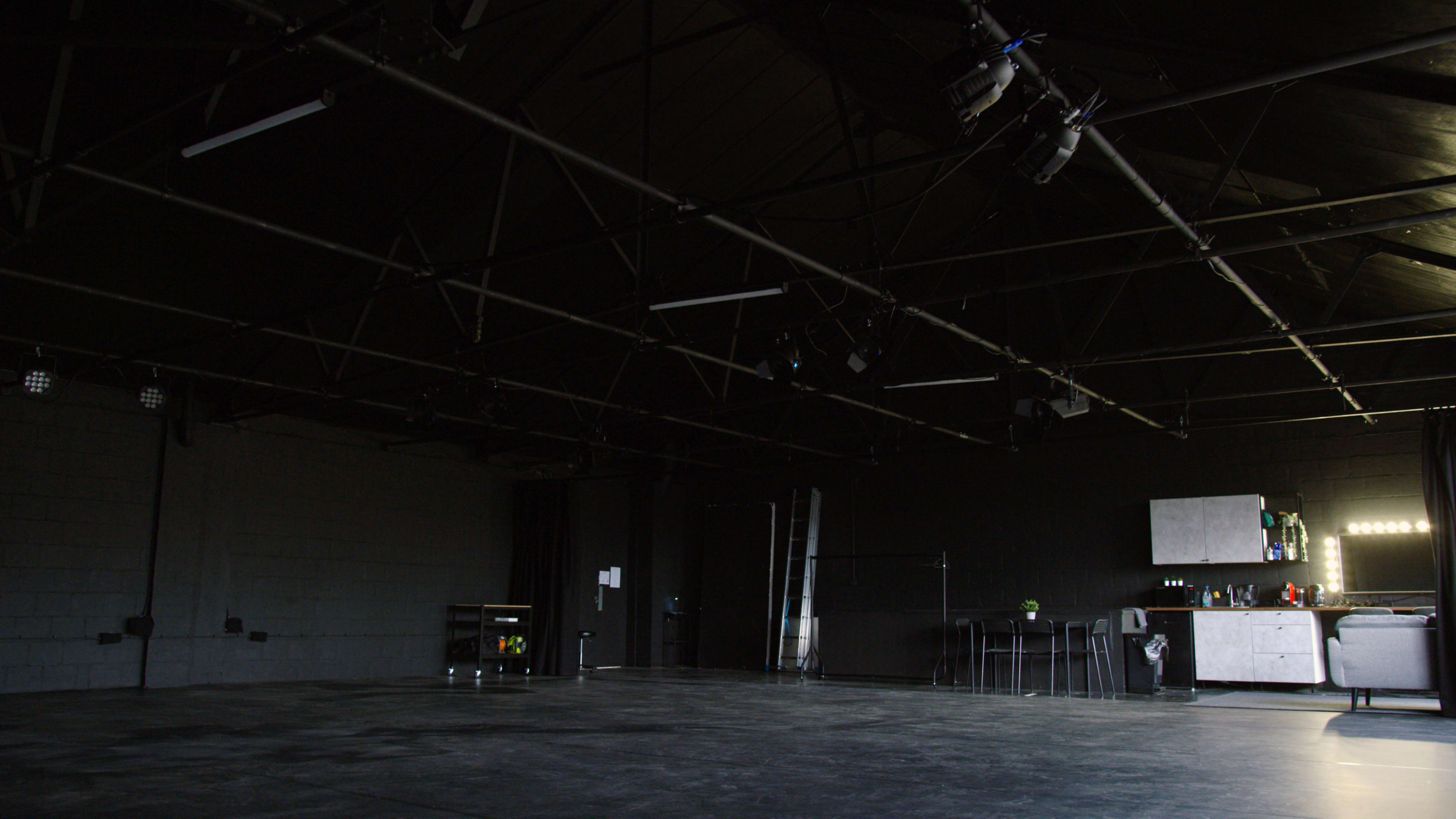A large, empty rehearsal space with a black ceiling, exposed beams, and a minimal kitchenette in one corner.