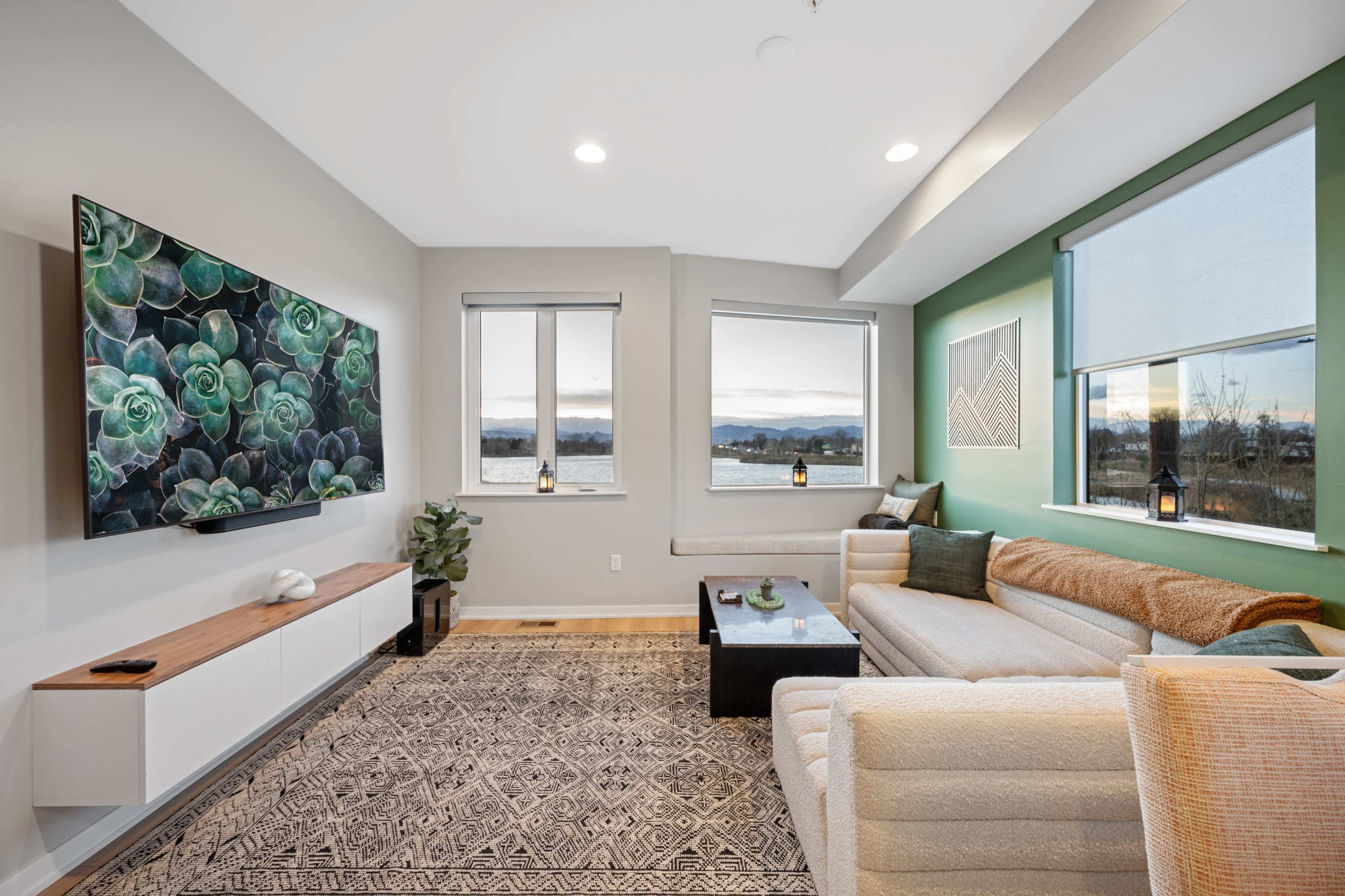 A modern living room features a large TV mounted on a wall, a beige sectional sofa, a coffee table, and windows with a view of the water.