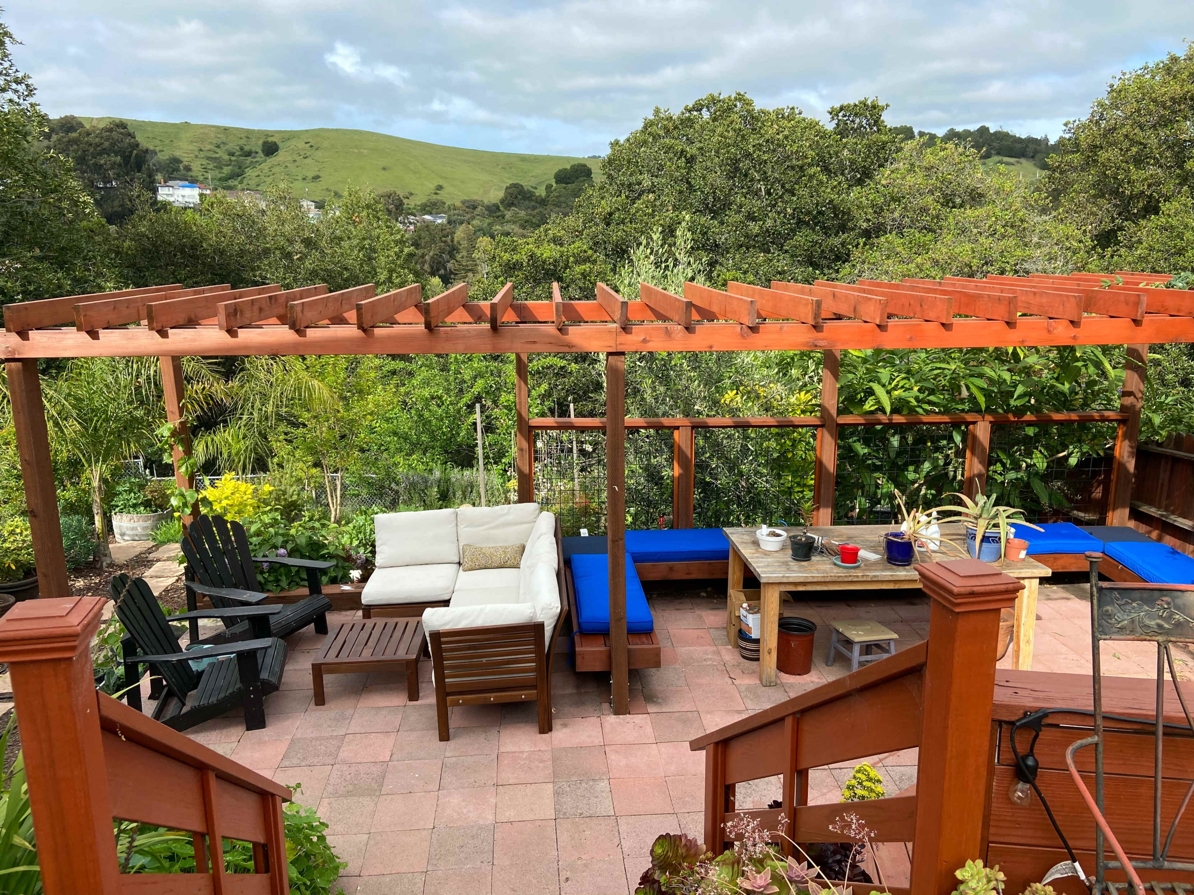 A backyard garden with a patio featuring a wooden pergola, outdoor seating, and potted plants surrounded by greenery and hills in the background.