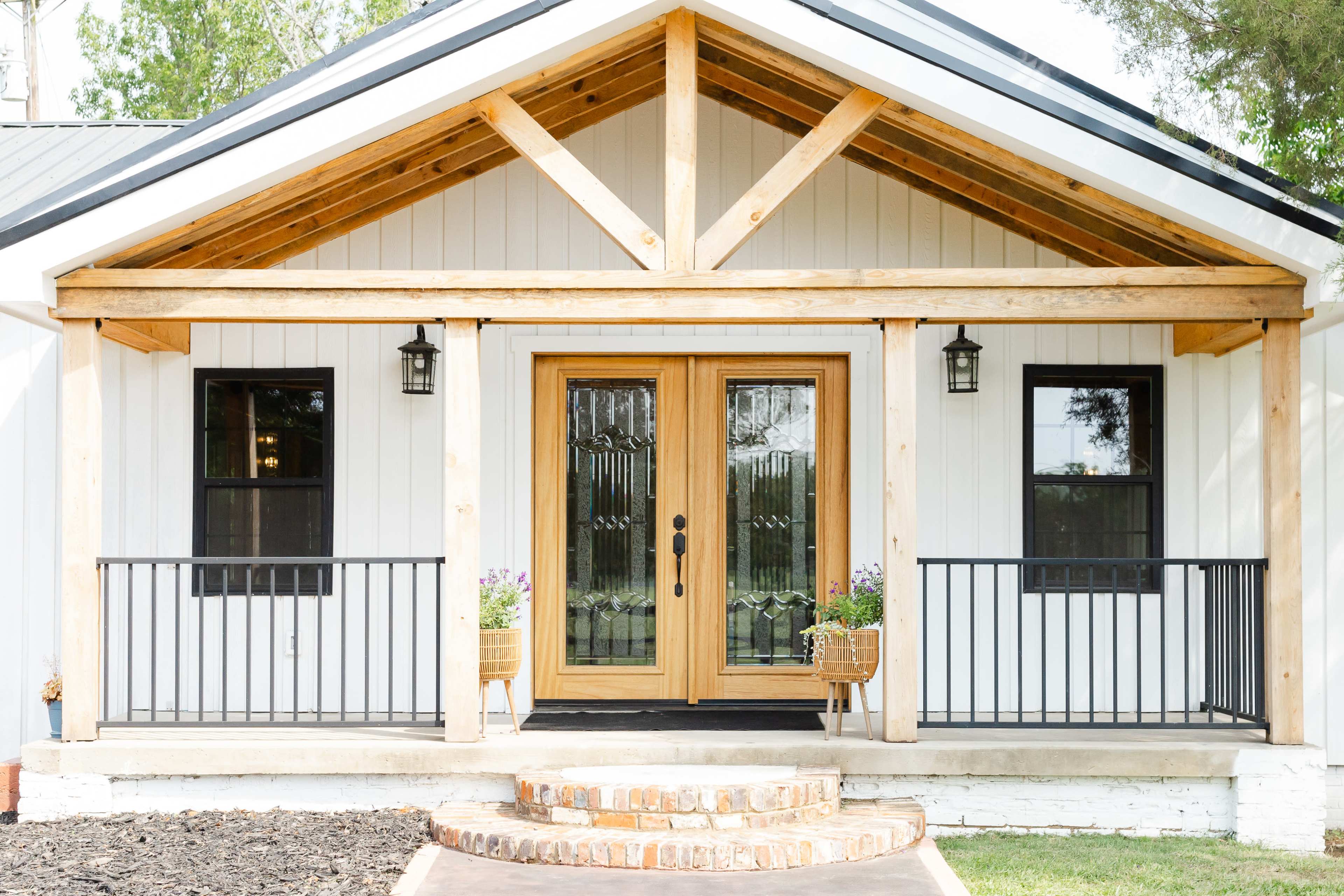 The image shows a house with a wooden porch featuring double doors, black railings, and two planters on either side, surrounded by greenery.