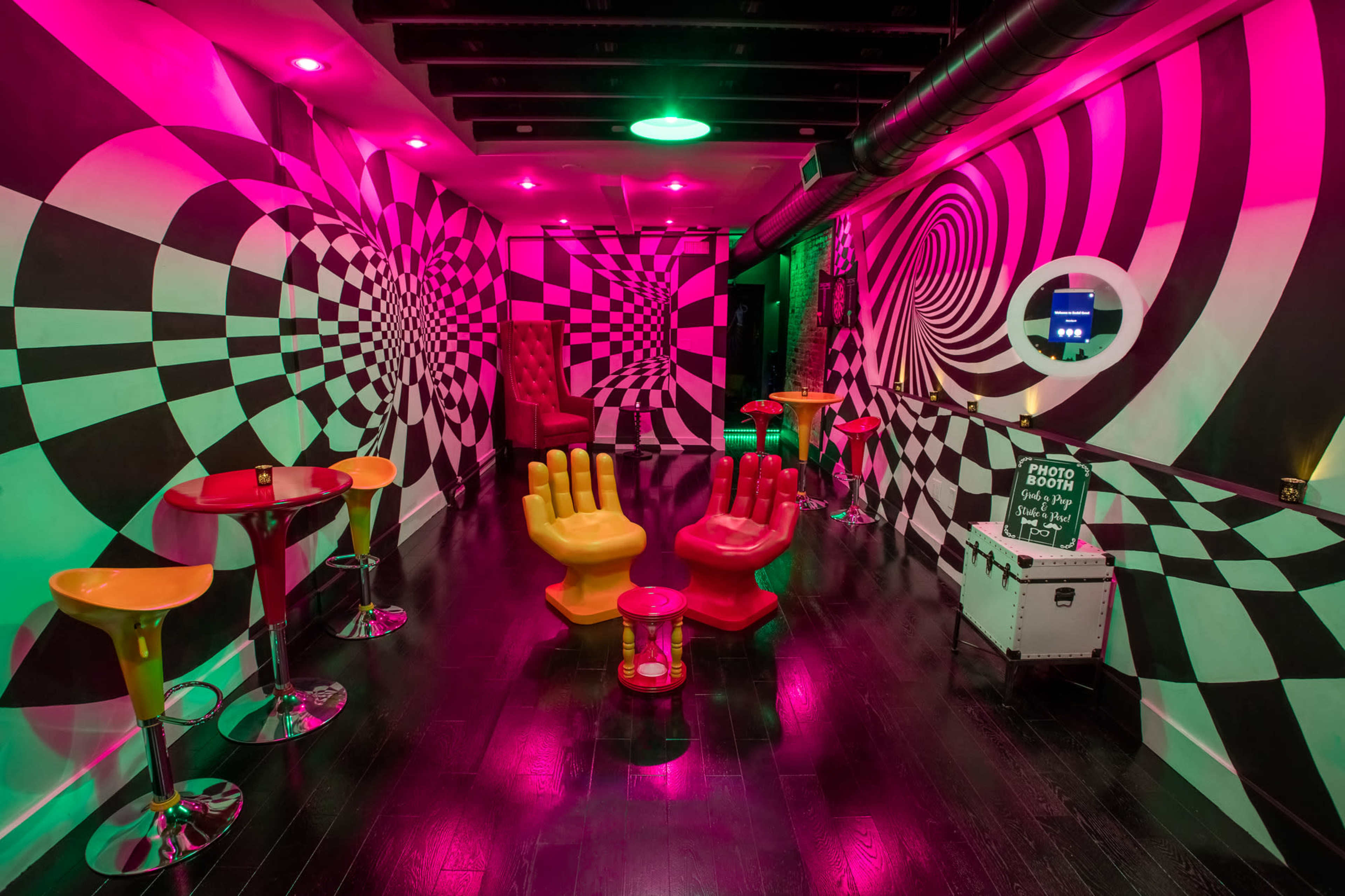 A room with a black and white checkered pattern and swirling designs on the walls, featuring colorful furniture including two large hand-shaped chairs and small round tables.